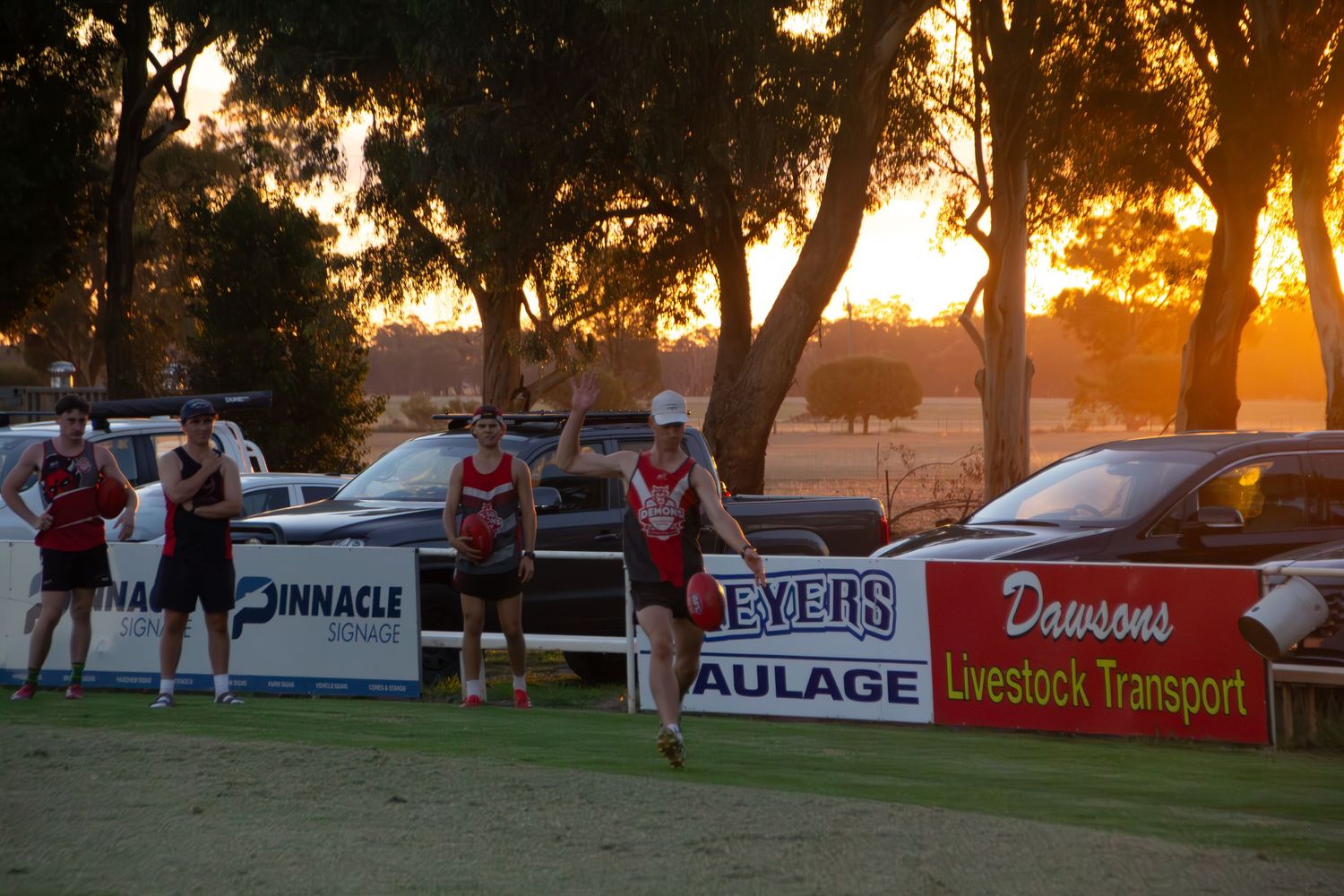 Athletes compete at sunset on an oval with sponsor banners including Dawsons Livestock Transport in regional Australia.