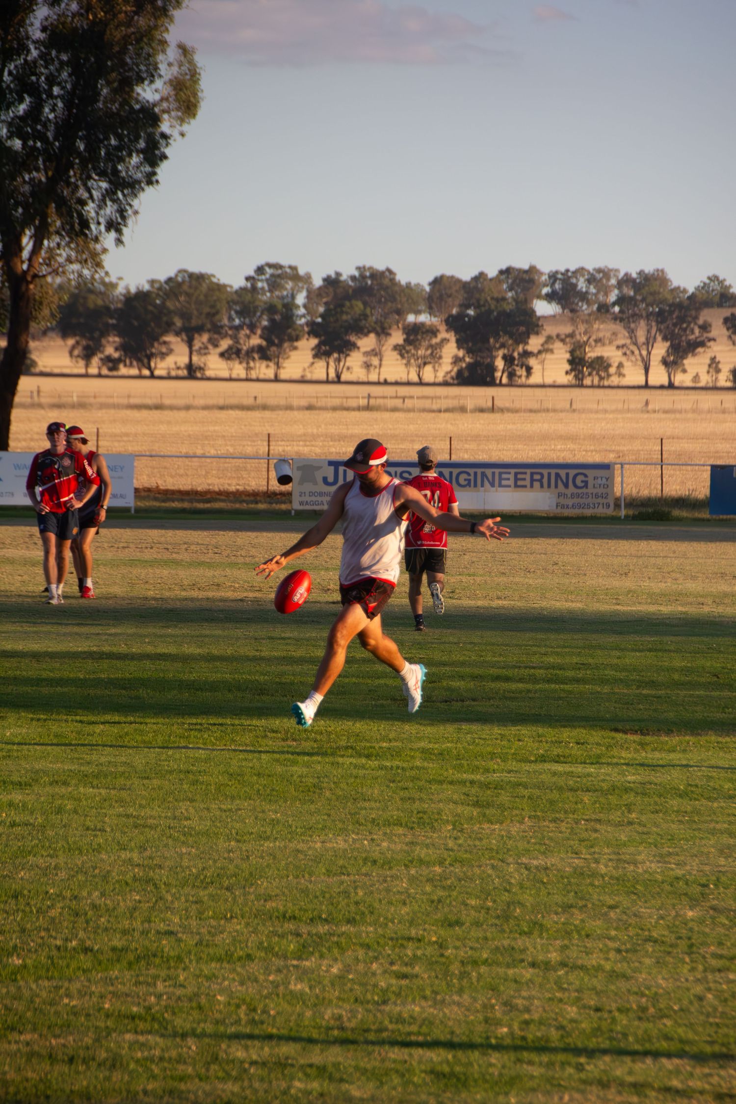 Soccer player in white jersey kicking ball on grass field during golden hour with teammates and trees in background.