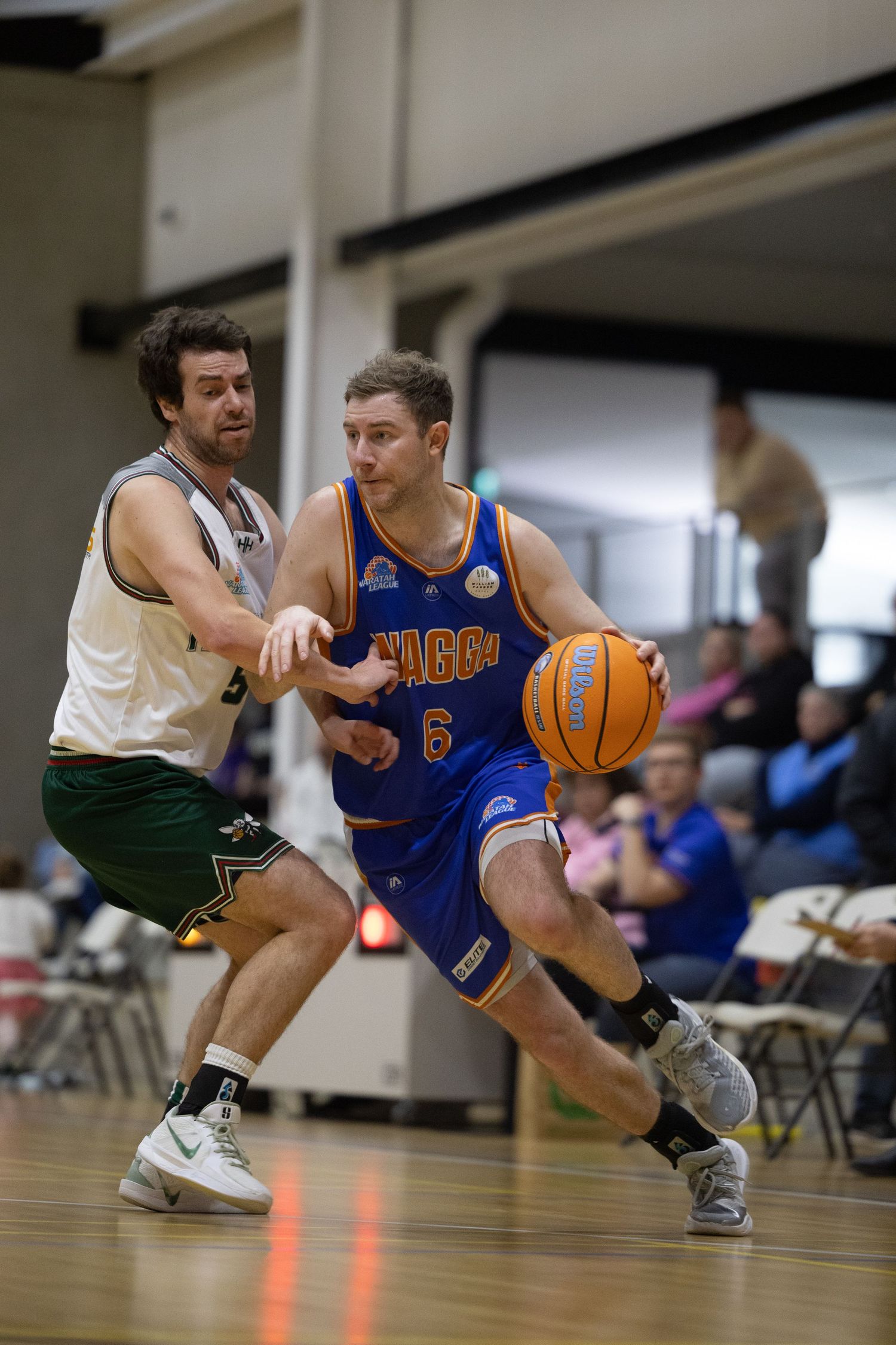 Basketball player in blue jersey dribbling past green-uniformed defender during indoor league game.