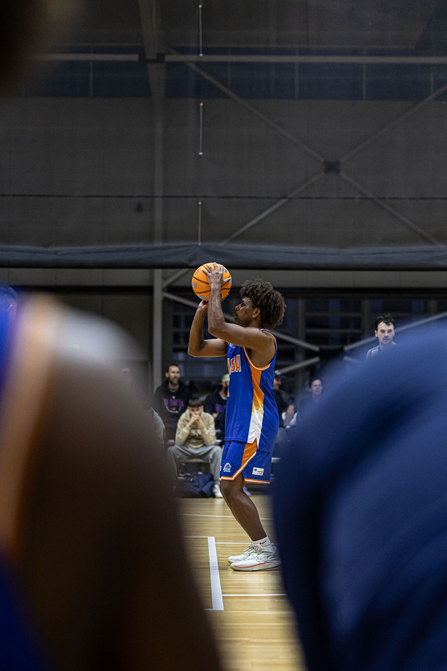 Basketball player in blue and orange uniform shoots the ball during an indoor game with spectators in background.