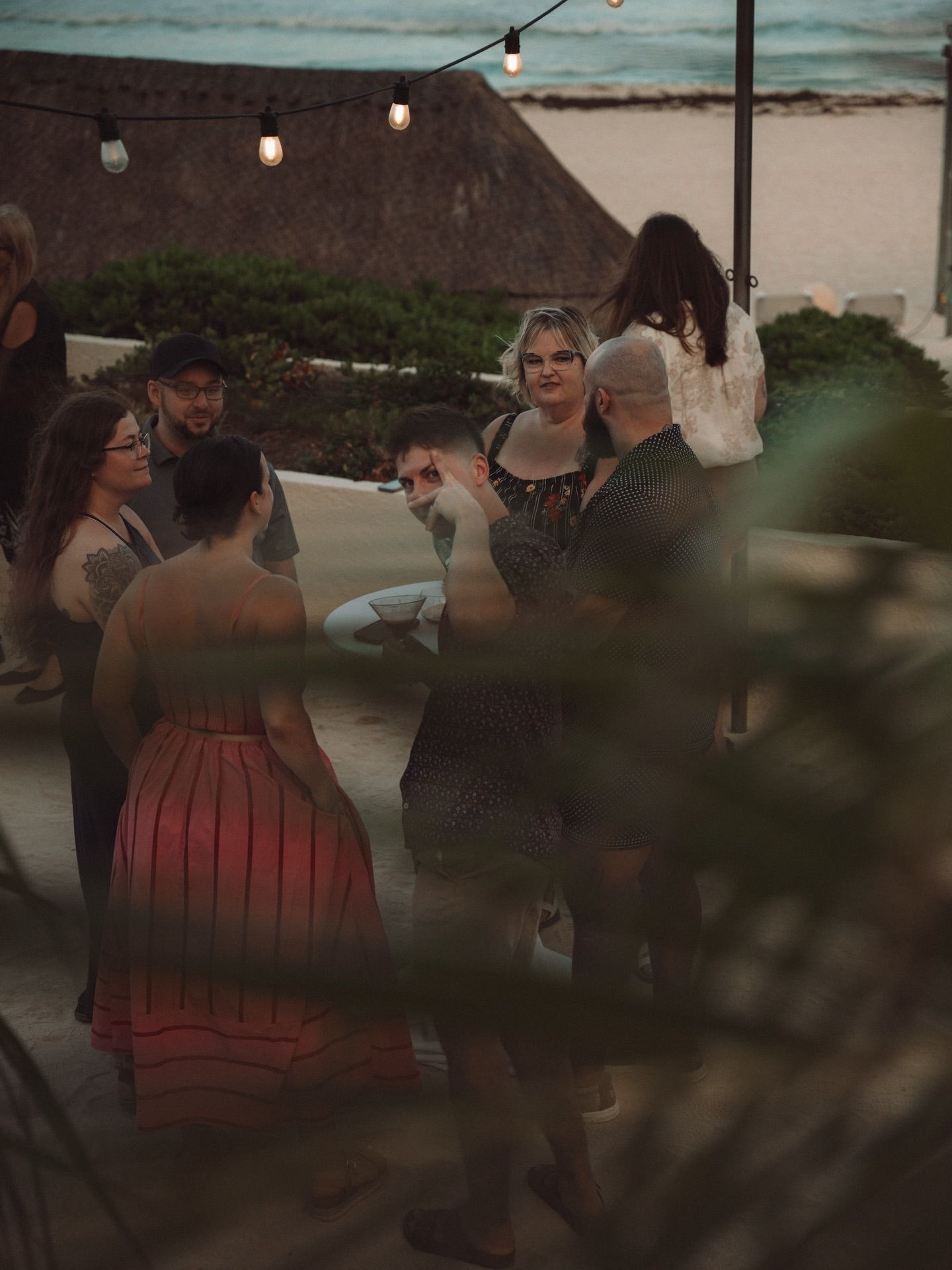 People gathered at an outdoor evening event with string lights and a beachy coastal backdrop.