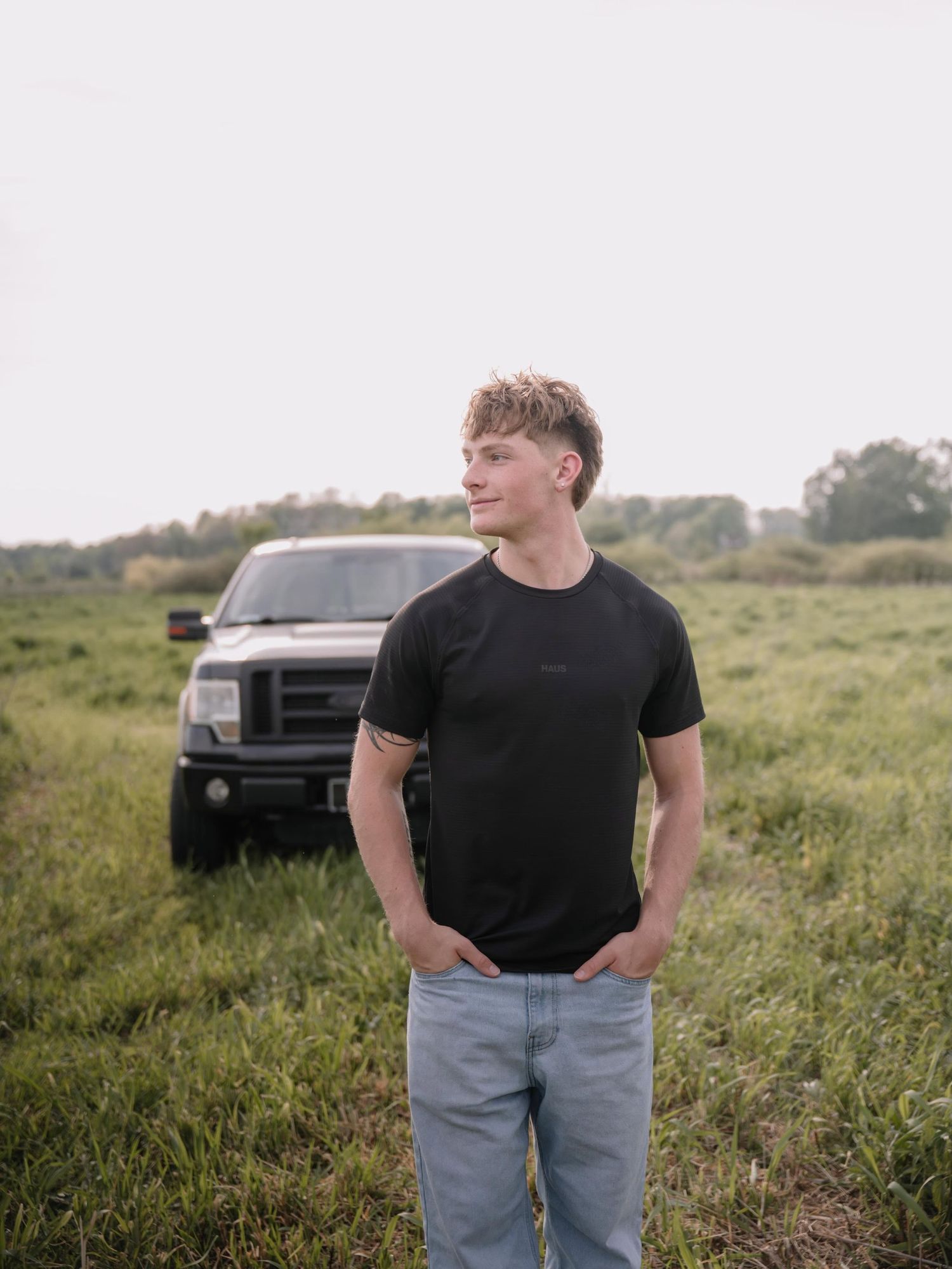 Young man in black t-shirt and jeans stands in green field with black pickup truck behind him.
