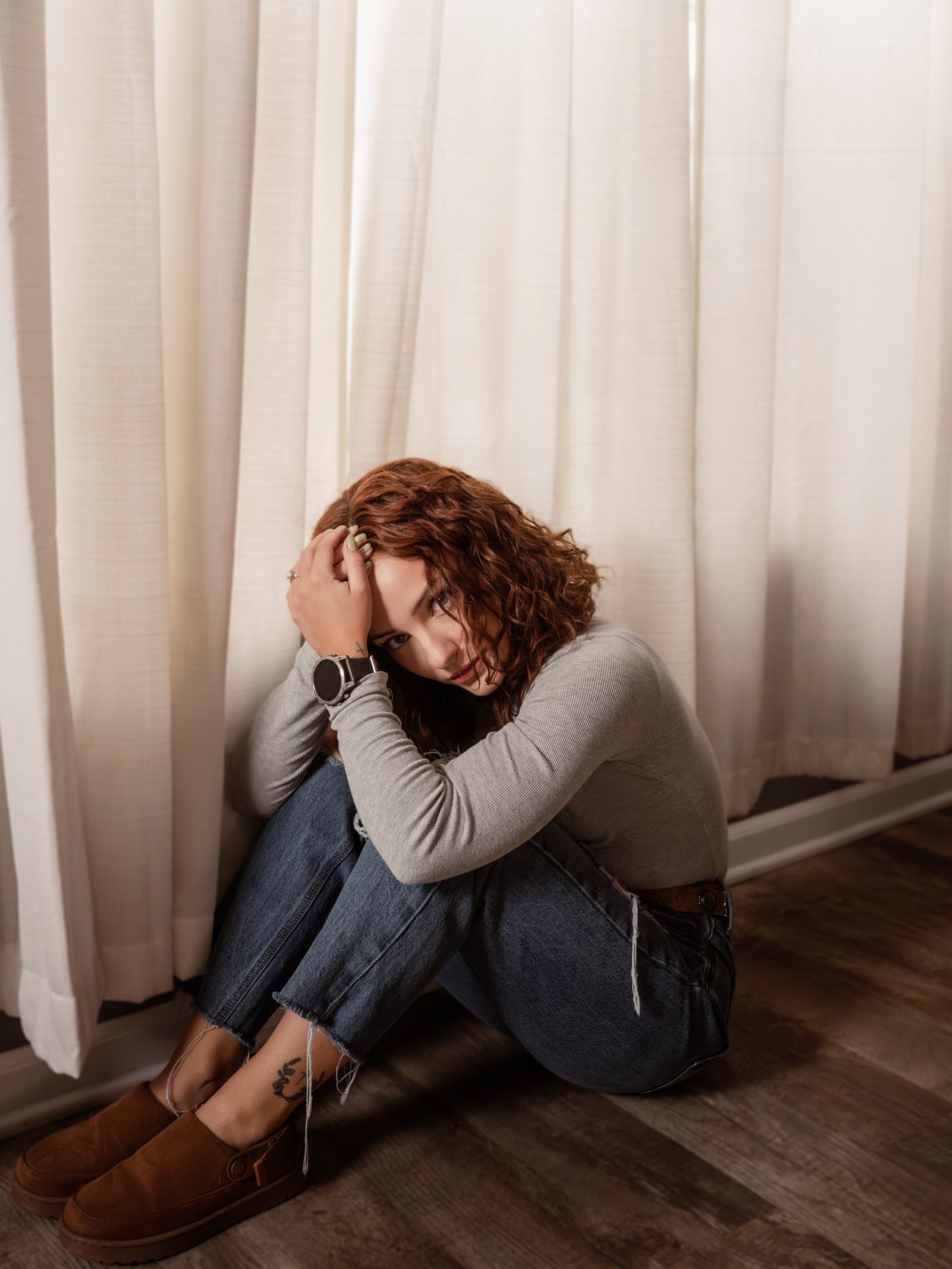 Person in grey sweater and jeans curled up against white curtained wall.
