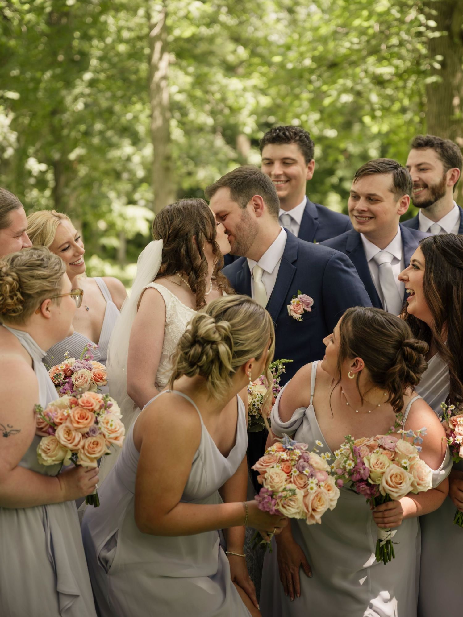 Wedding party gathered outdoors as bride and groom share a kiss, surrounded by smiling bridesmaids in gray dresses holding floral bouquets.
