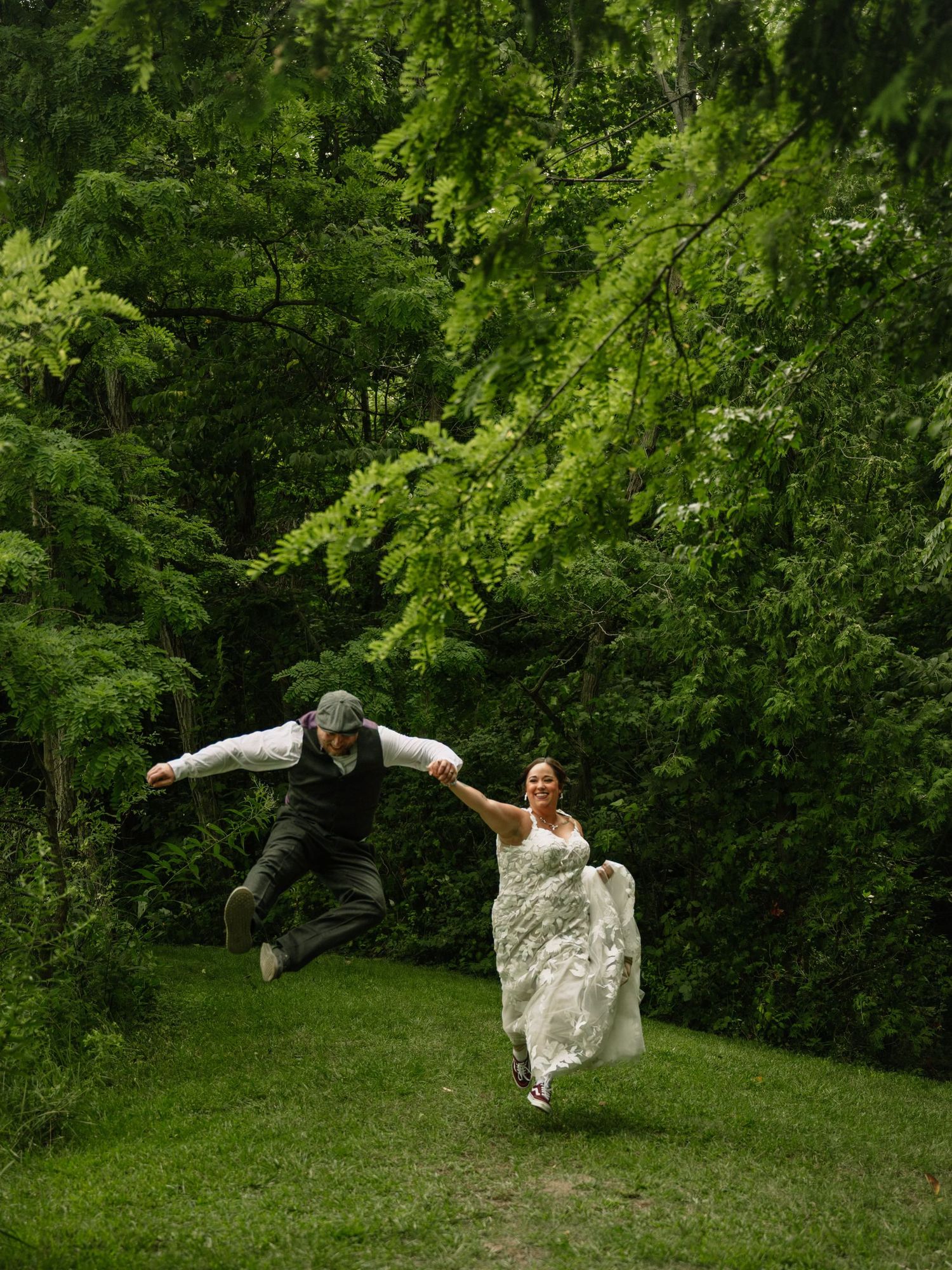 Joyful bride and groom jumping and laughing together in a lush green forest setting during their wedding day.