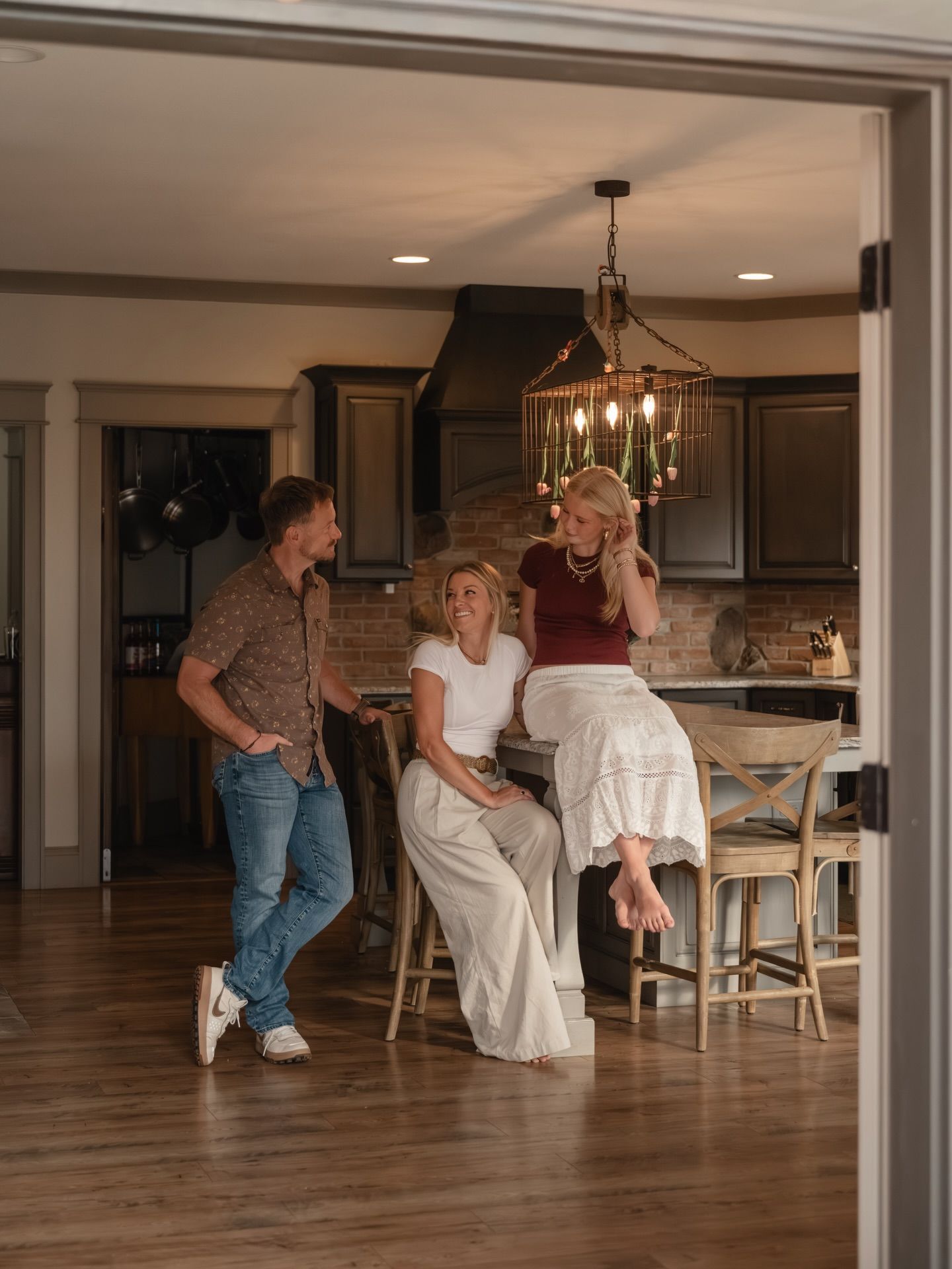 Three people socializing in a modern kitchen with dark cabinets, hardwood floors, and a rustic chandelier.
