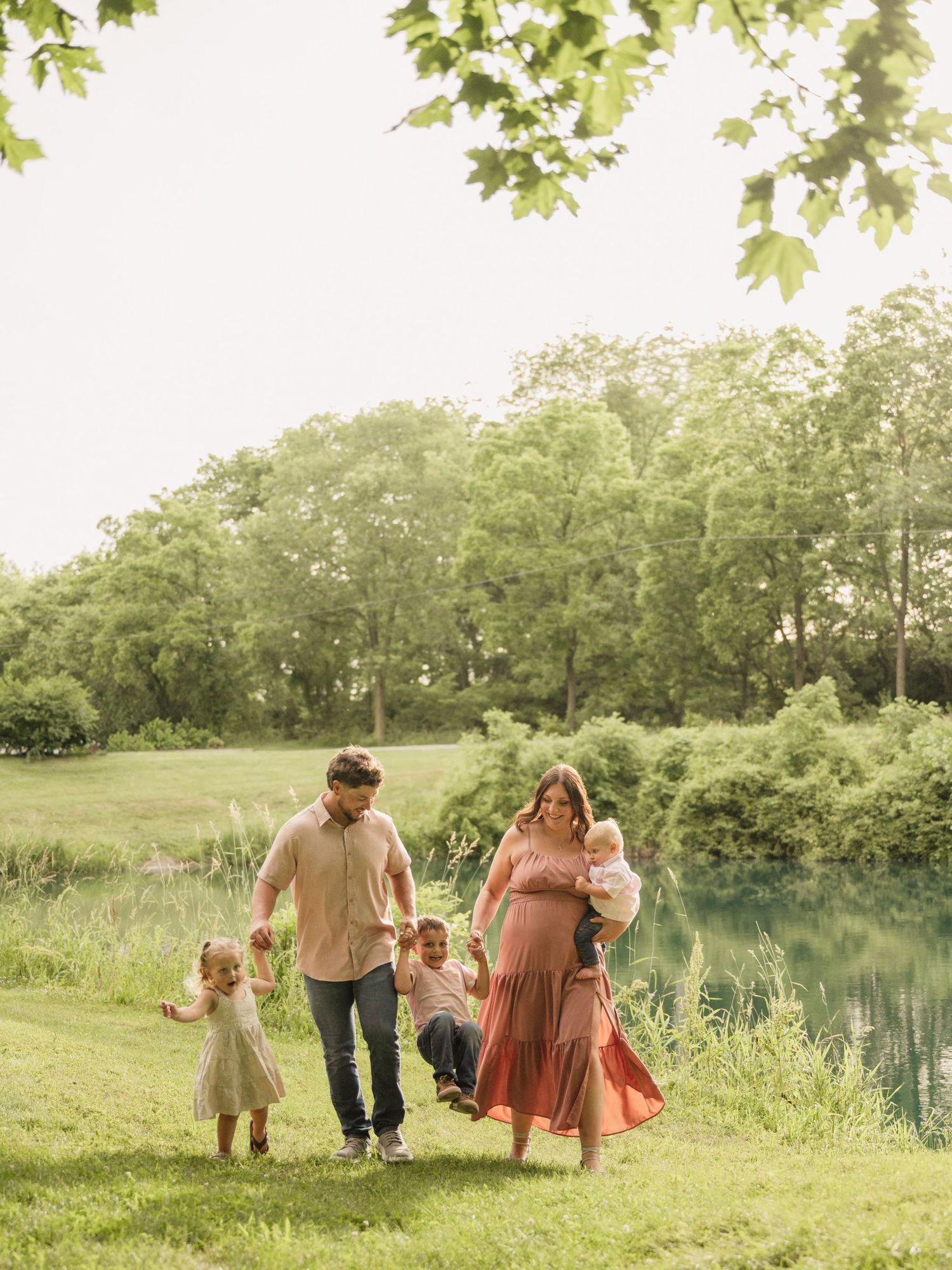 A family walks by a pond in a lush green park, with parents swinging a child between them on a sunny day.