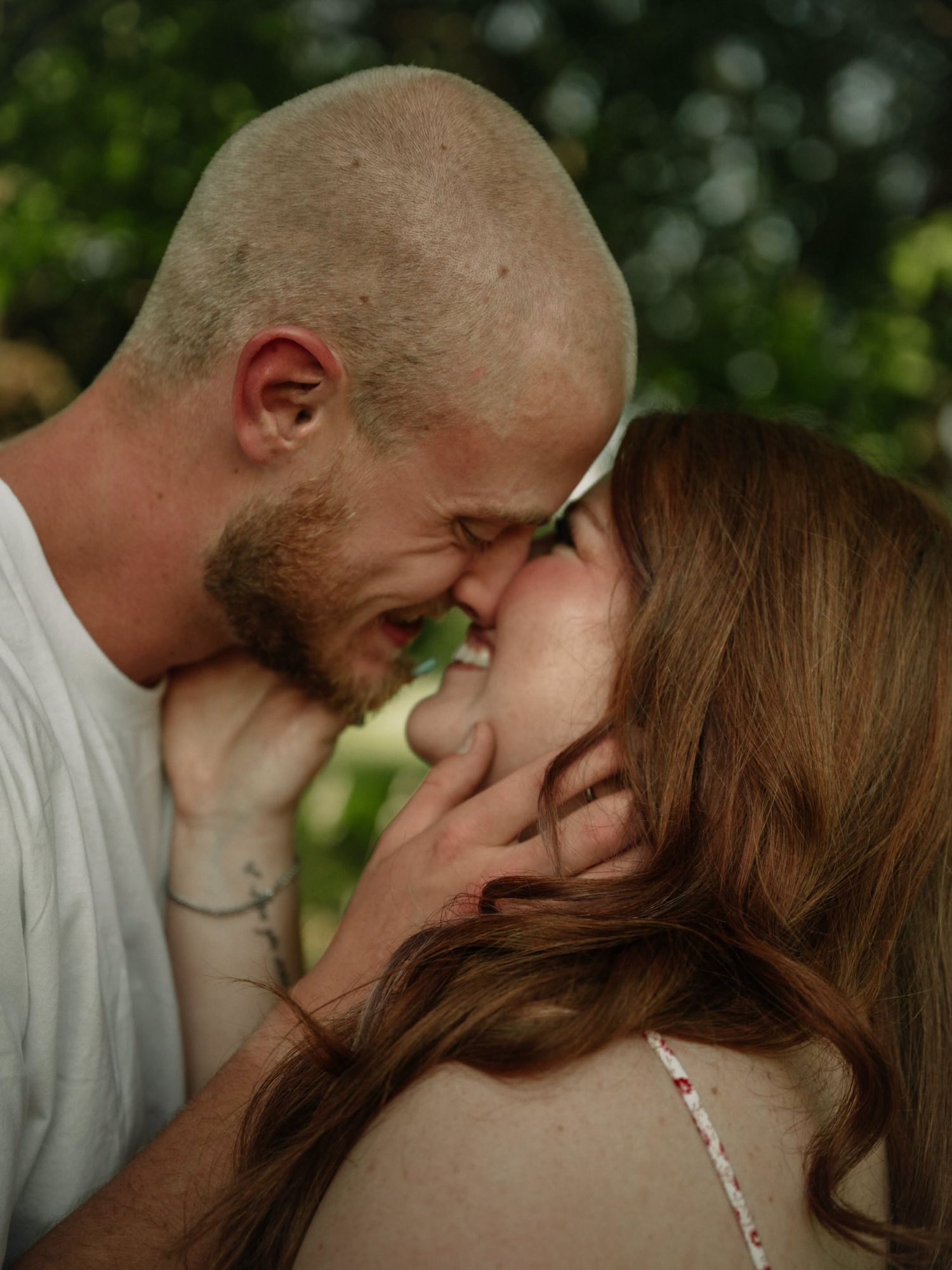 A couple sharing an intimate moment, foreheads touching and almost kissing outdoors with green foliage background.