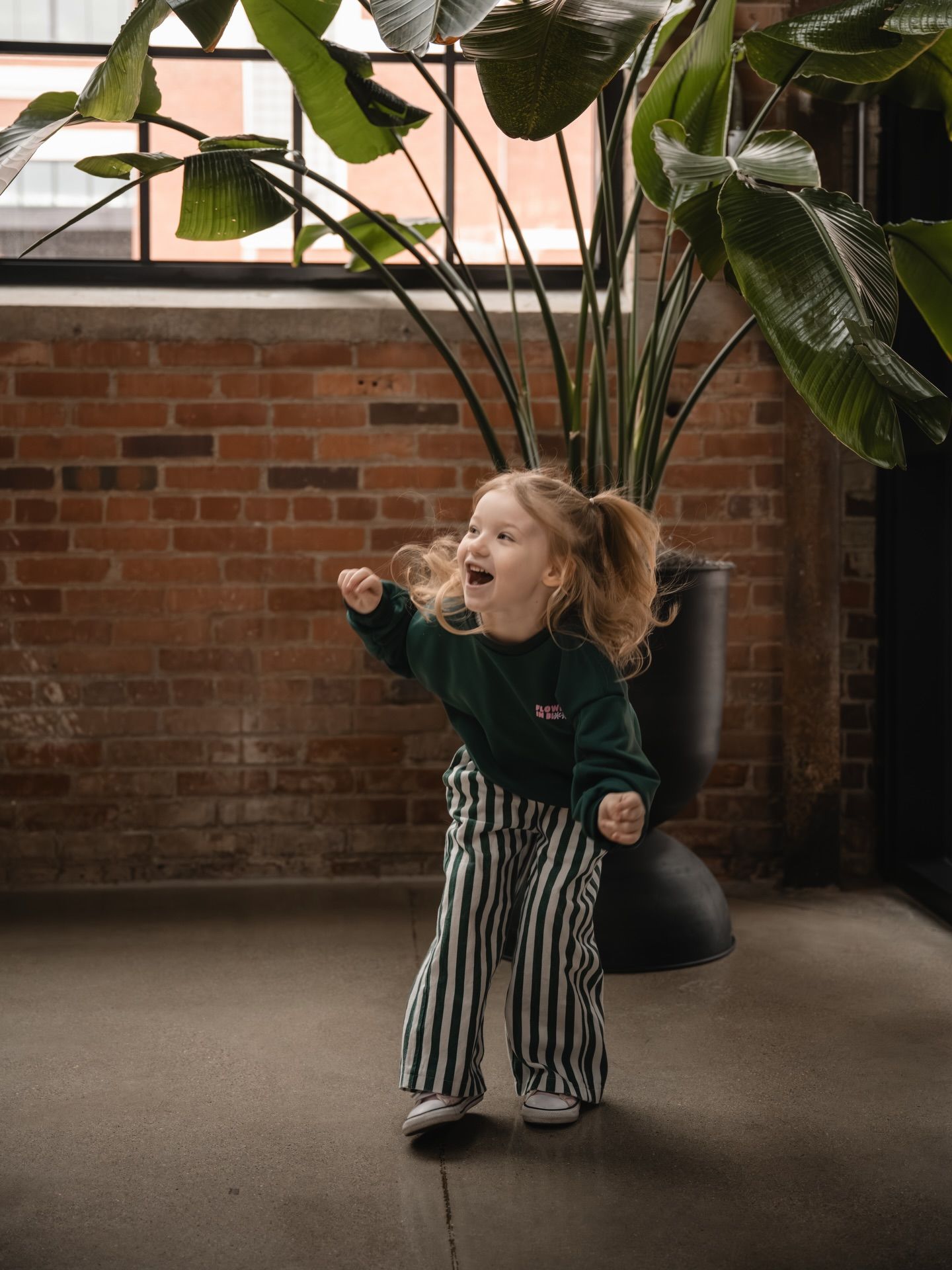 A joyful toddler in striped pants and green sweater dances energetically indoors near a large plant and brick wall.