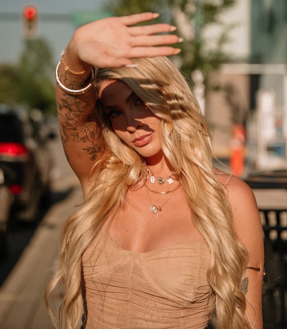 Blonde woman in beige dress shielding sun with tattooed arm, wearing layered necklaces on urban street.