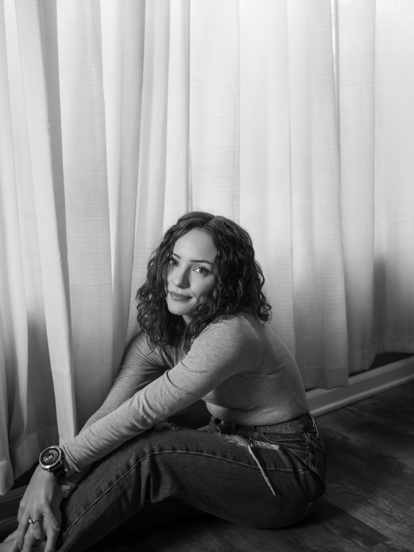 A black and white portrait of someone sitting by a window with sheer curtains.