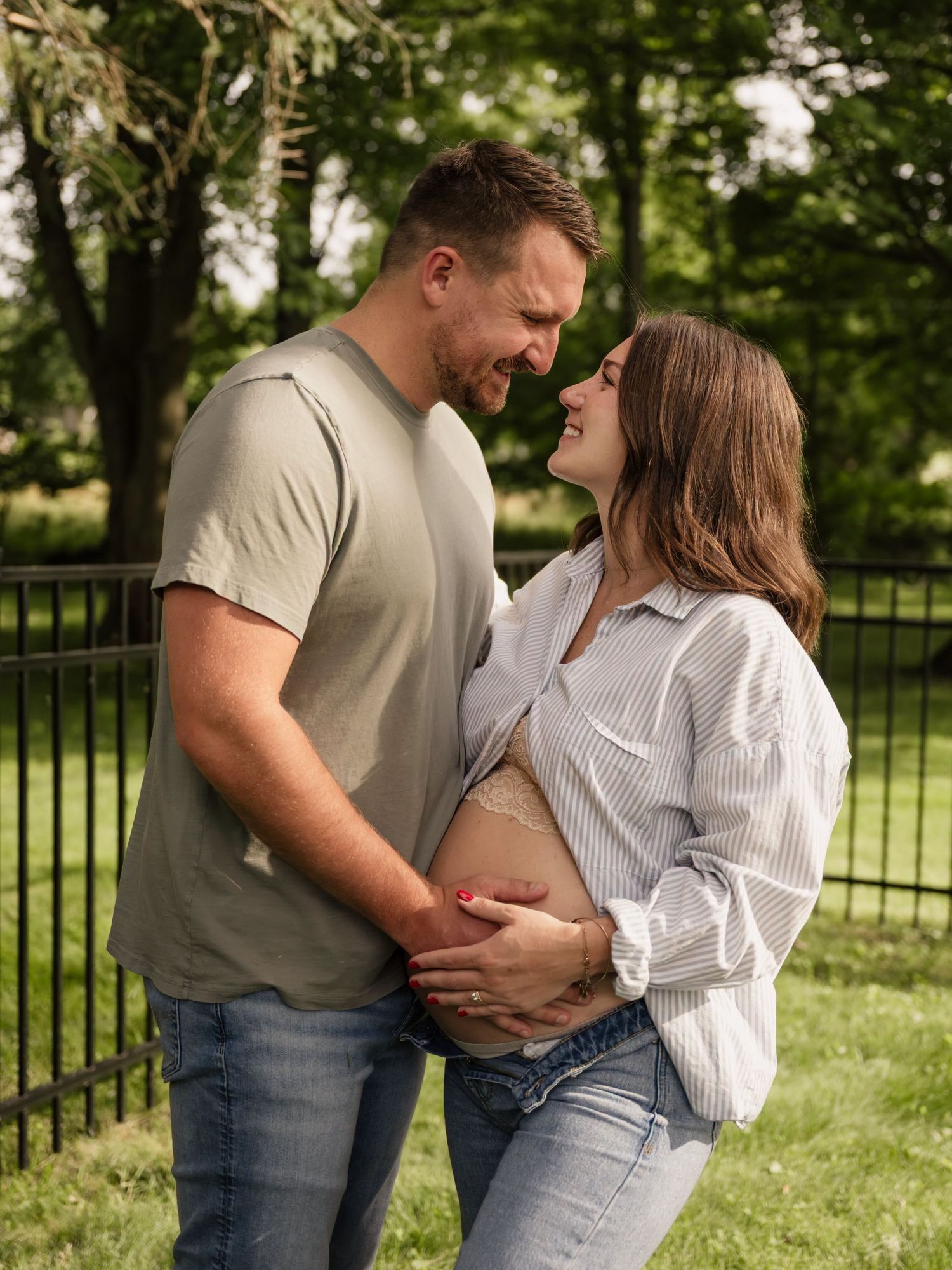 A couple shares a tender moment outdoors, cradling a pregnant belly, surrounded by lush green trees and a black fence.