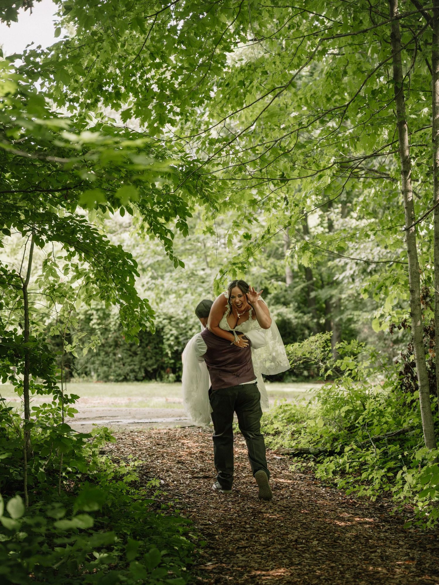 A groom carries his bride through a lush green forest path on their wedding day.