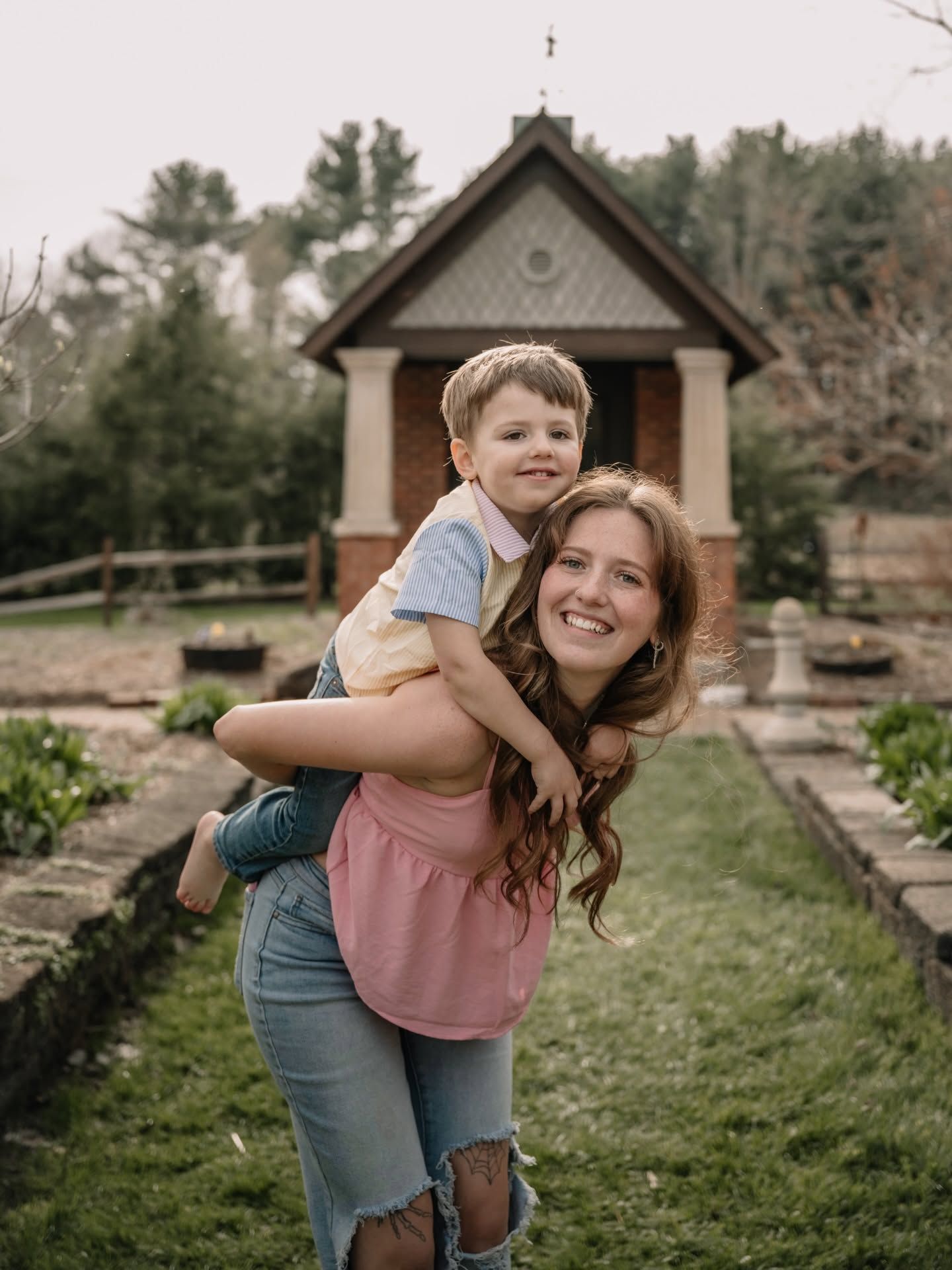 A smiling woman gives a young boy a piggyback ride in a garden with a small brick chapel in the background.