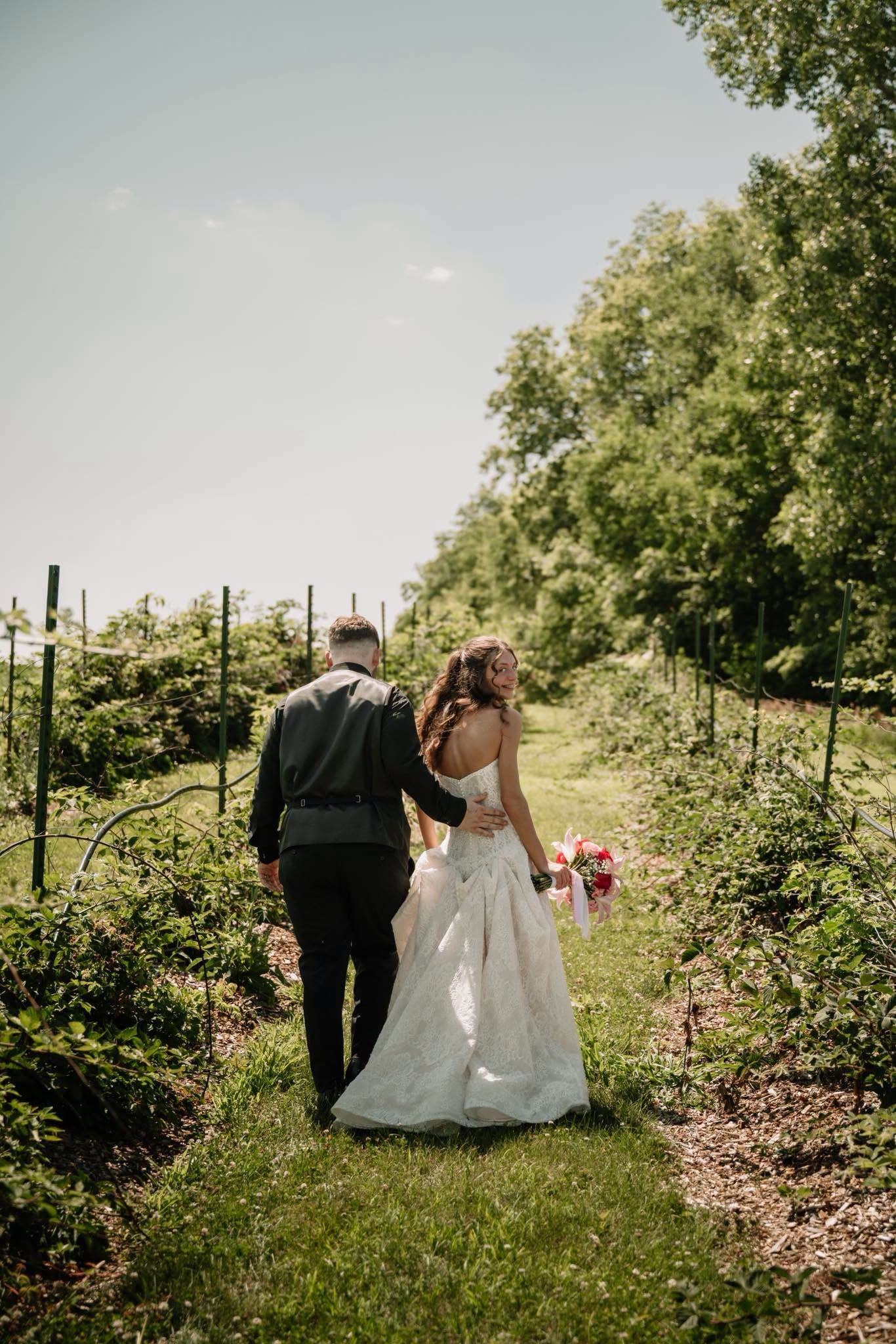 A bride and groom walk hand in hand through a lush vineyard path on their wedding day.