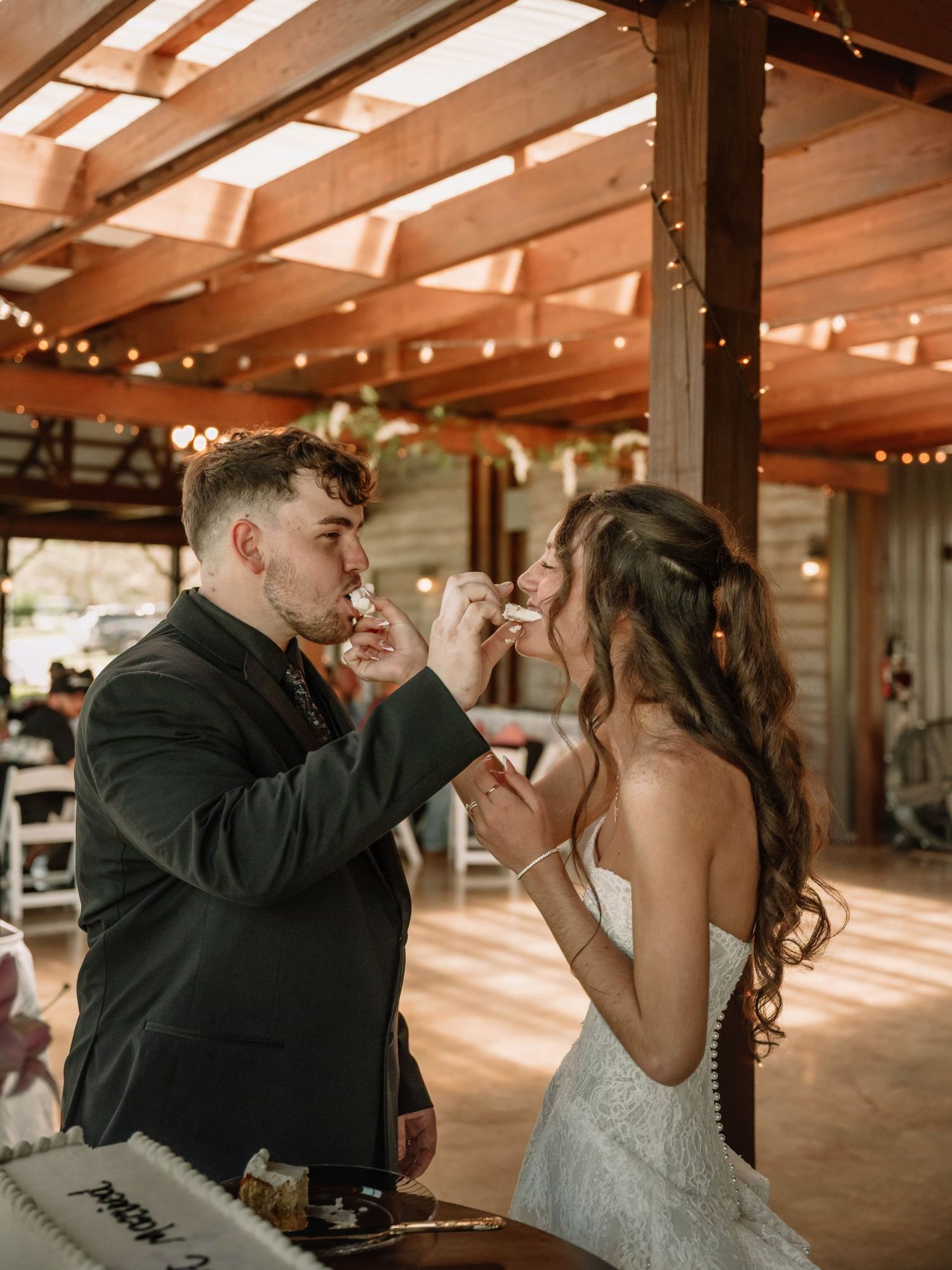 Bride and groom feeding each other wedding cake under a rustic pergola with string lights at their reception.