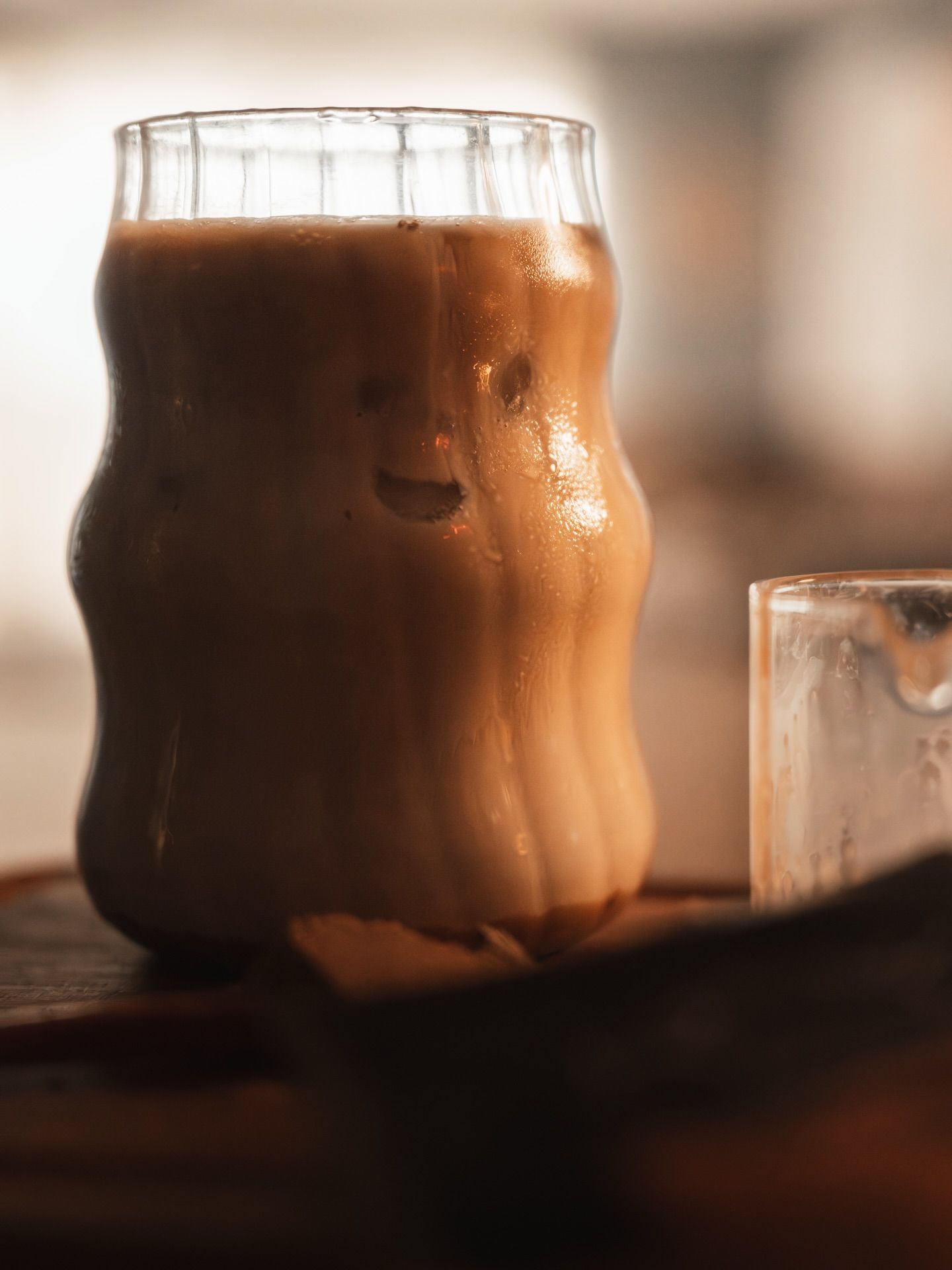 A wavy glass filled with creamy iced coffee sits on a wooden surface beside a small ice-filled glass.