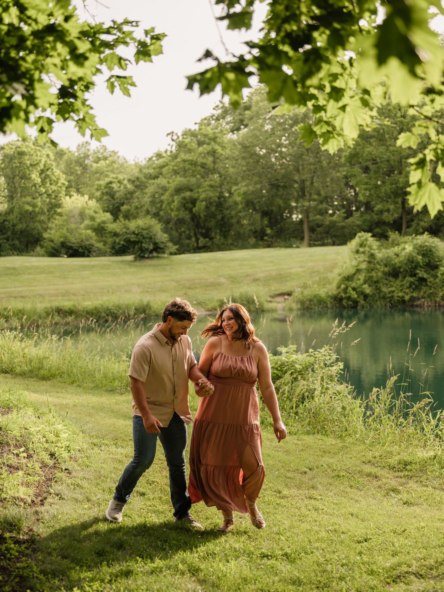 A couple walks hand in hand through a lush green park beside a tranquil pond on a sunny day.