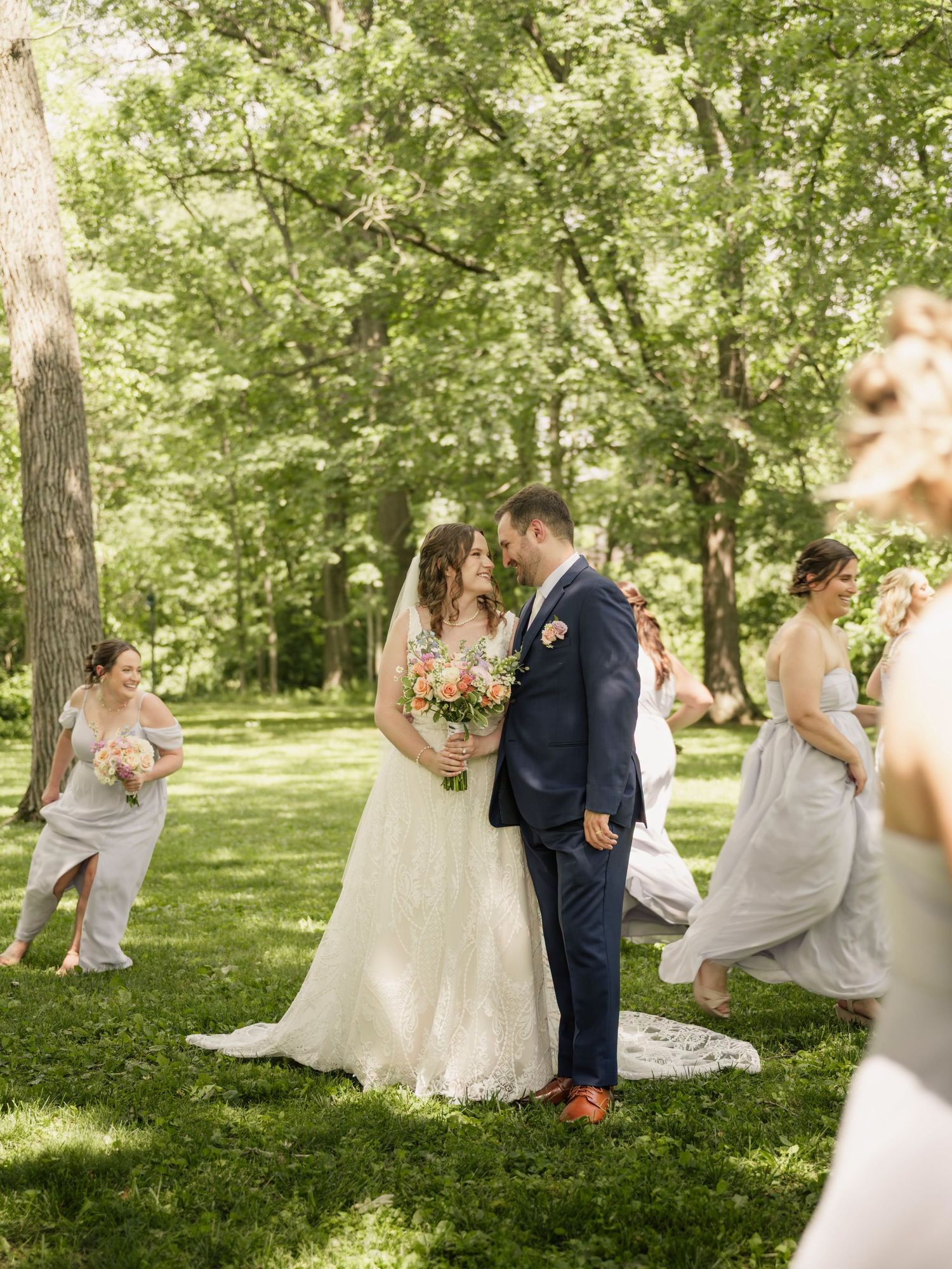 Bride and groom share a romantic moment outdoors surrounded by bridesmaids in gray dresses in a lush green garden.