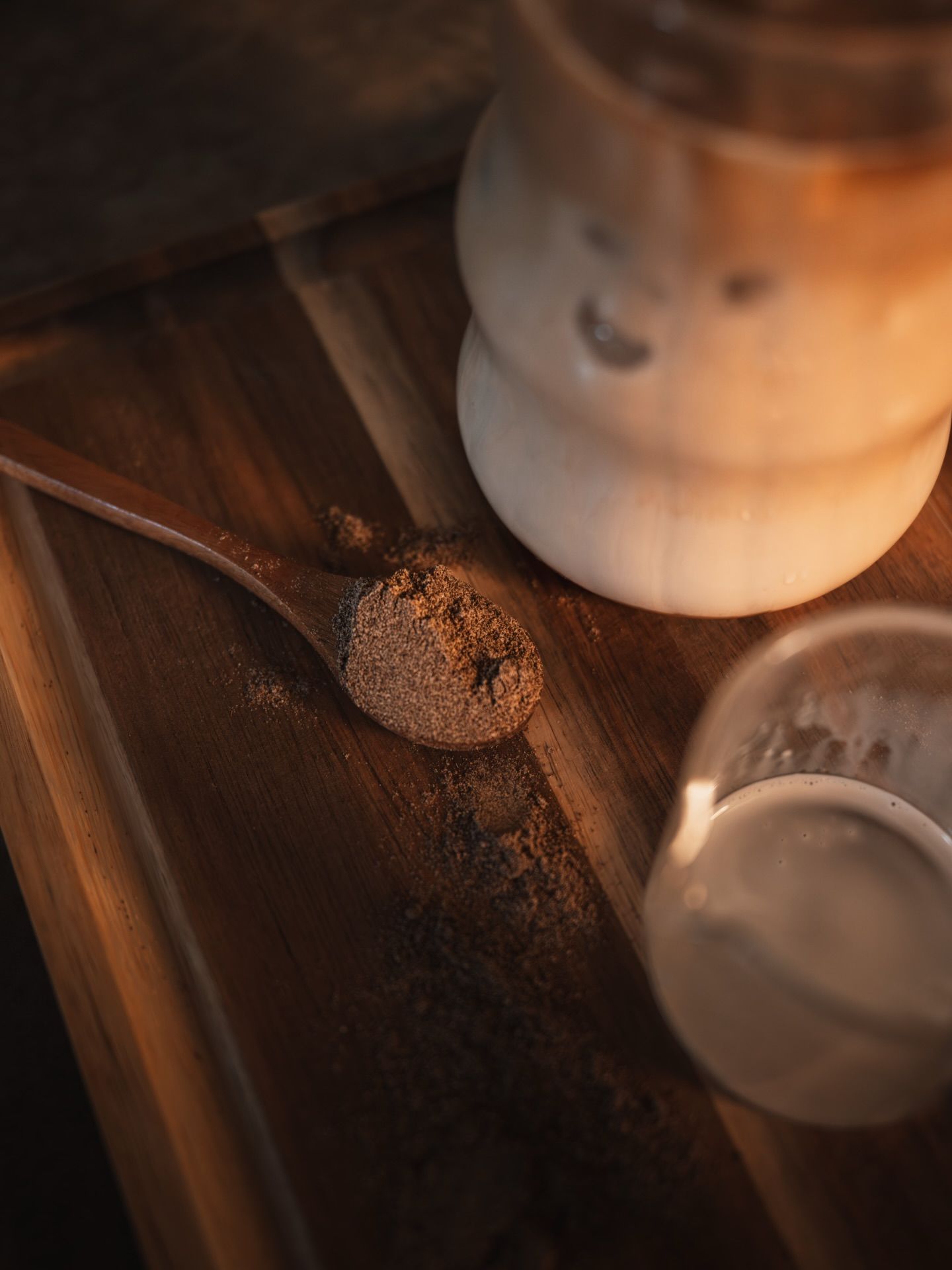 A spoonful of brown powder on a wooden tray beside a glass jar and cup in warm moody lighting.