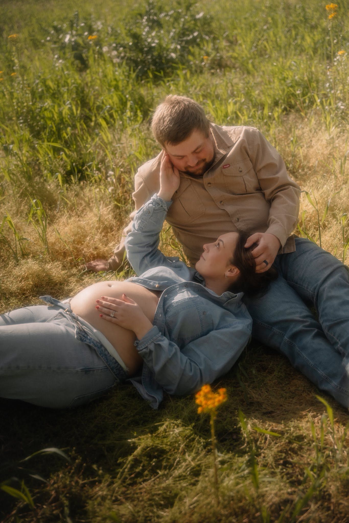 Pregnant couple relaxing in a sunny green field, woman lying on man's lap, sharing a tender moment outdoors.