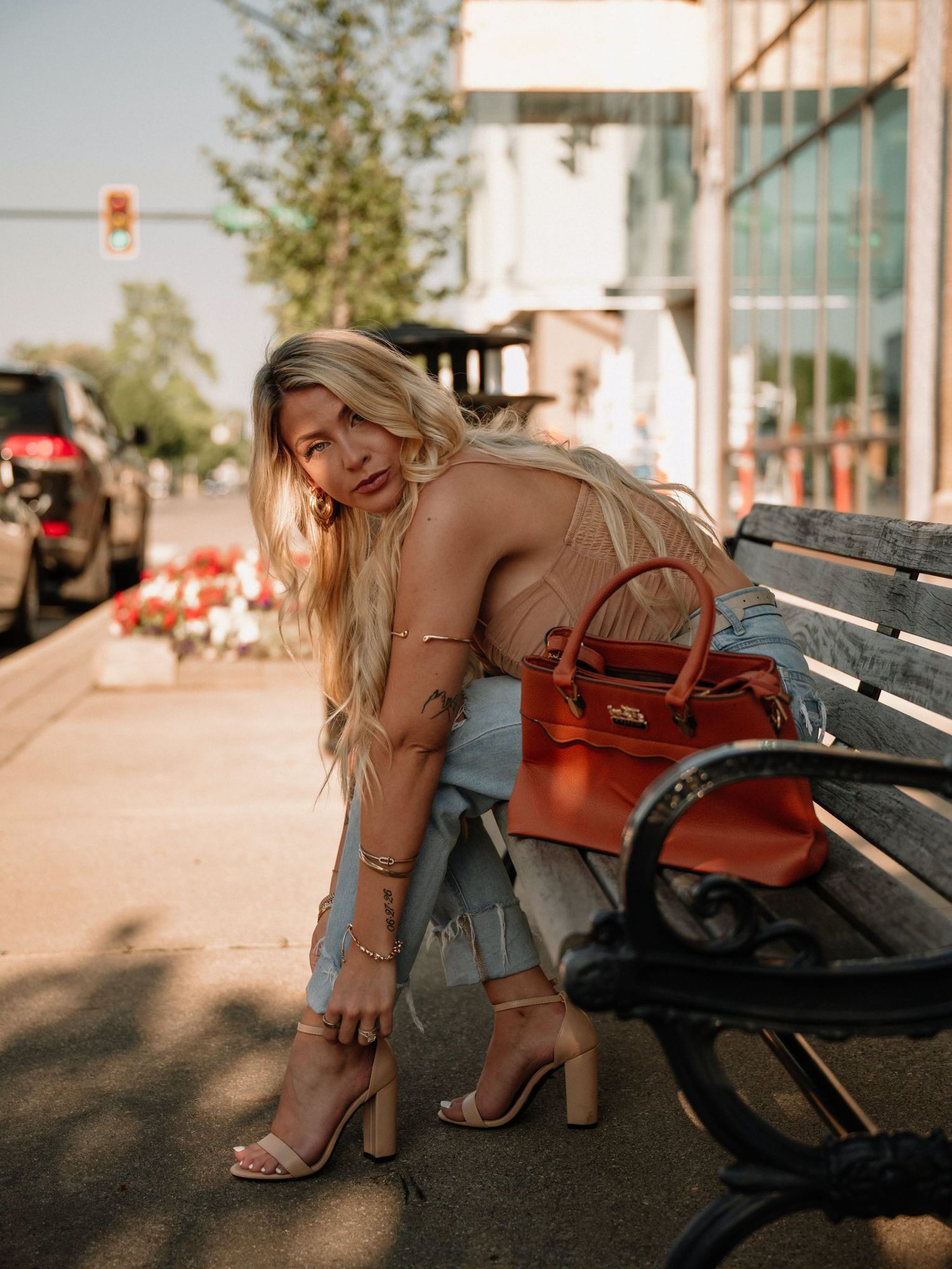 Blonde woman in beige top, jeans, and heels adjusting her shoe on a city bench with an orange handbag.