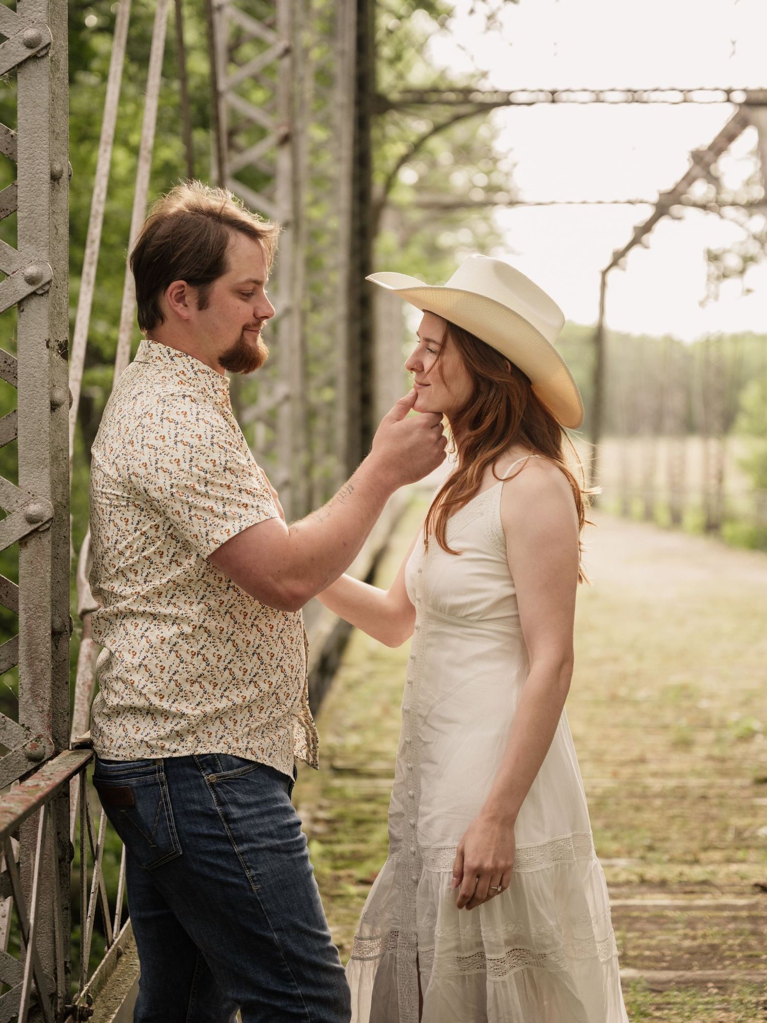 Couple sharing a tender moment outdoors, woman in white dress and cowboy hat, man in patterned shirt and jeans.