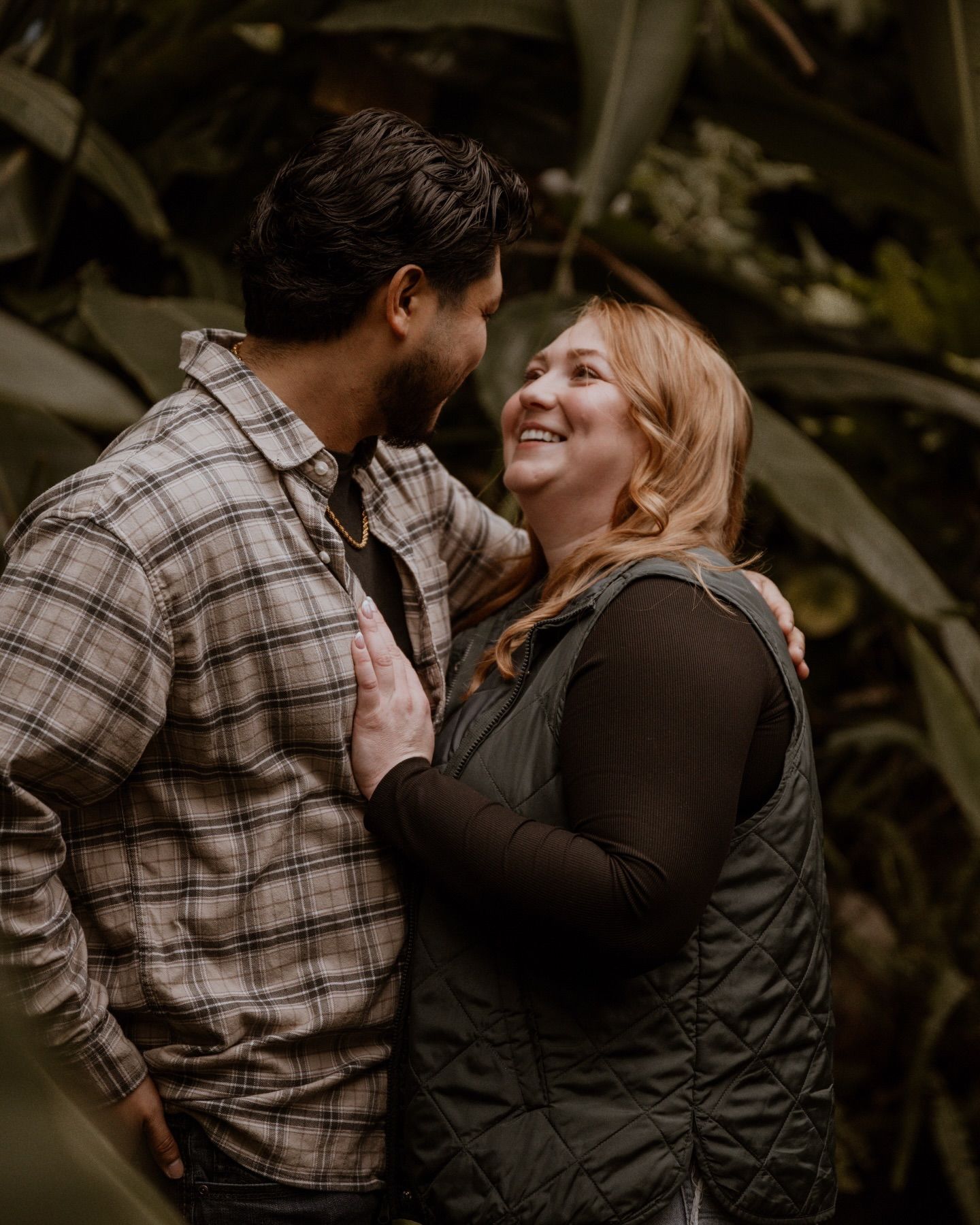 Romantic couple embraces in a moody outdoor setting wearing casual plaid and dark clothing.
