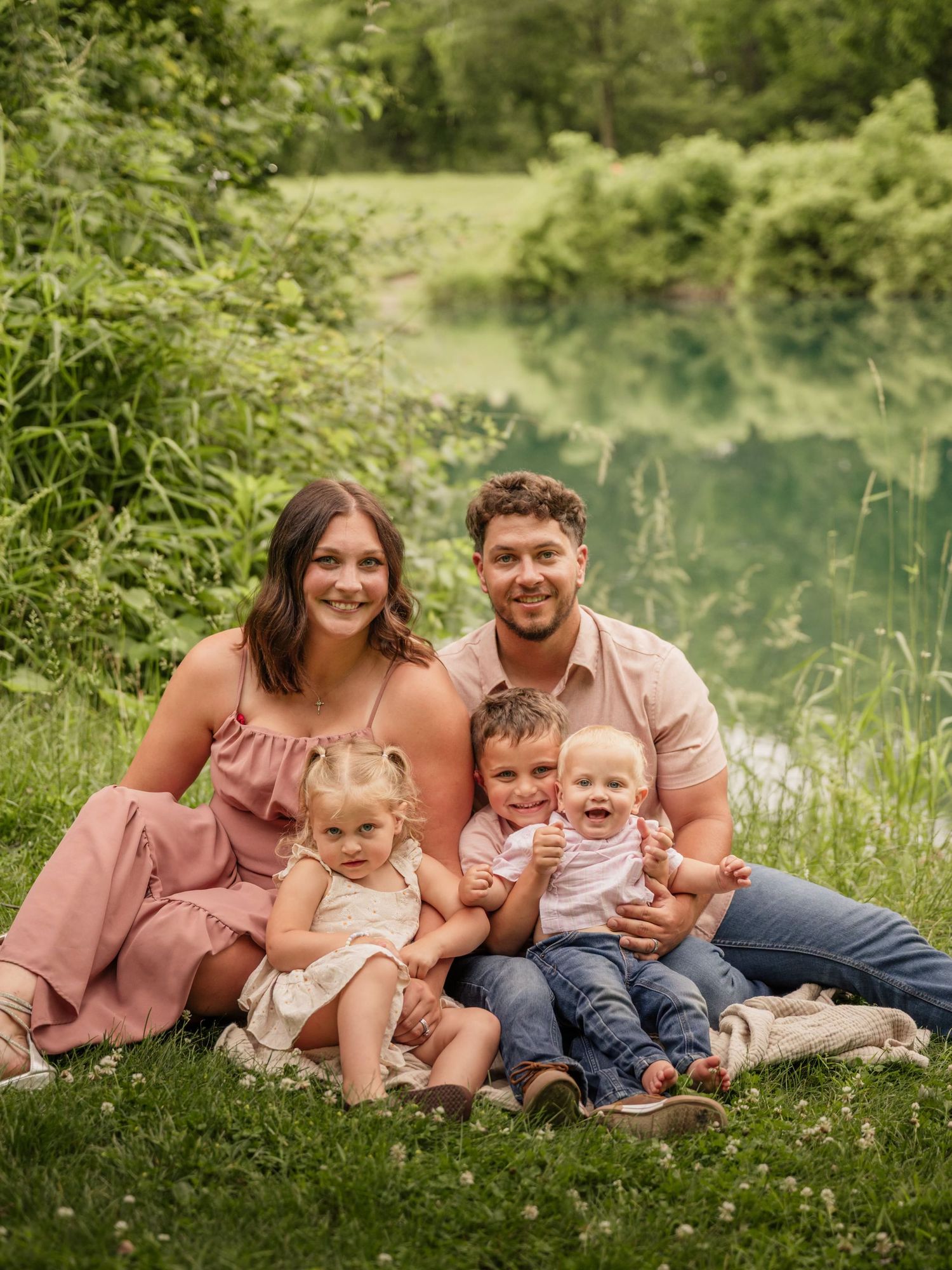 A happy family of five poses on green grass near a pond, wearing coordinating pink and neutral outfits.