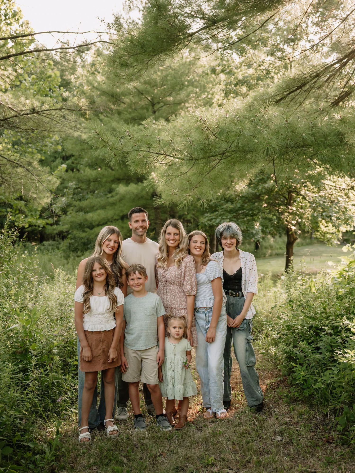 A joyful family of seven poses together outdoors surrounded by lush green trees and wildflowers.