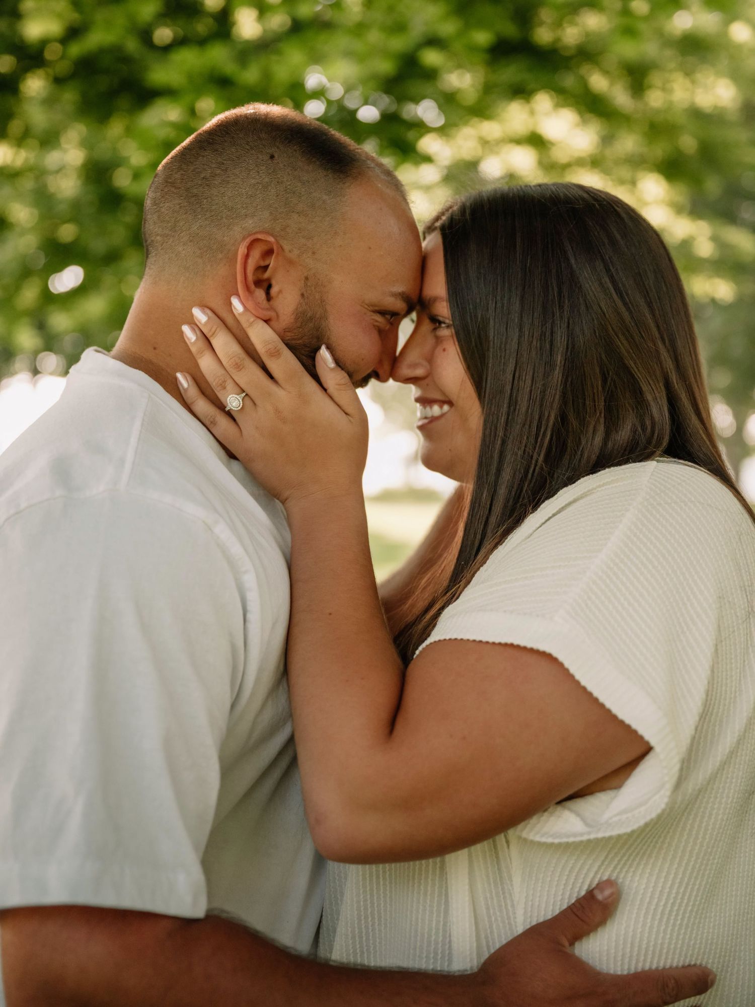 Couple in white outfits touching foreheads and smiling, woman wearing engagement ring, outdoors with green trees.