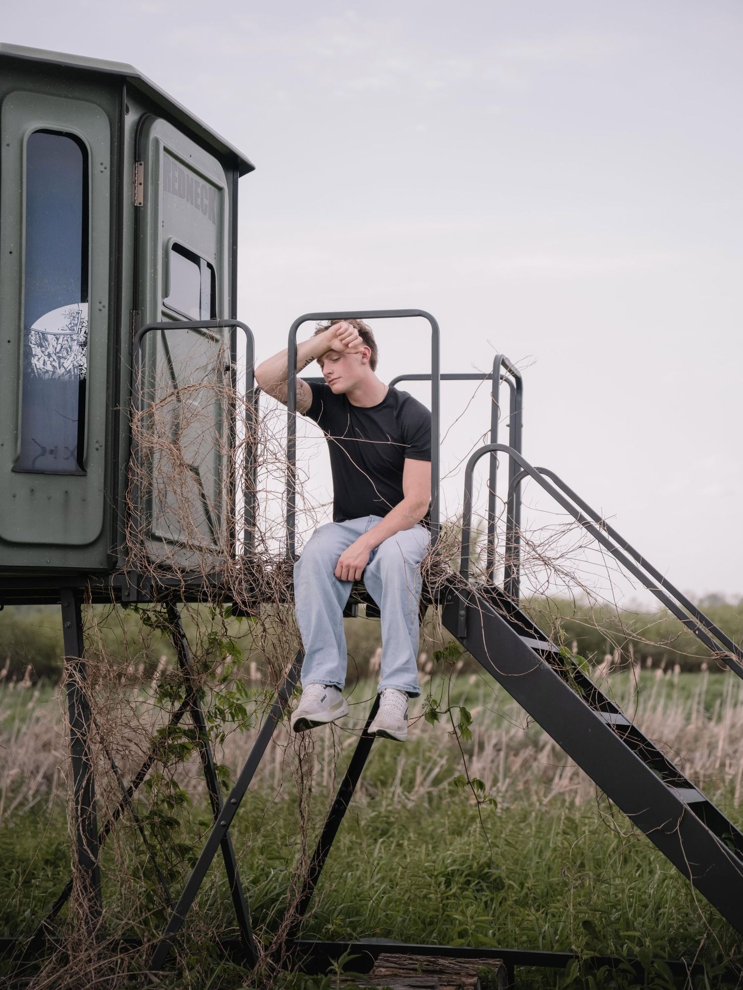 Young man in black t-shirt and jeans sitting on metal stairs of a hunting blind in a rural field.