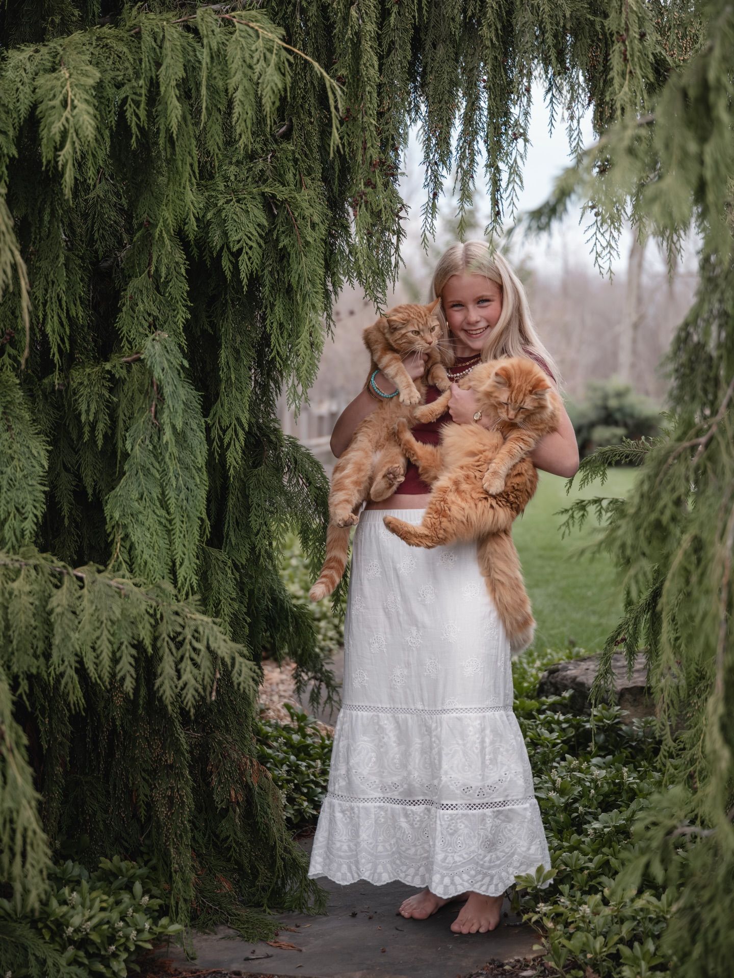 Blonde girl in white dress holding two orange tabby cats outdoors under weeping evergreen trees.