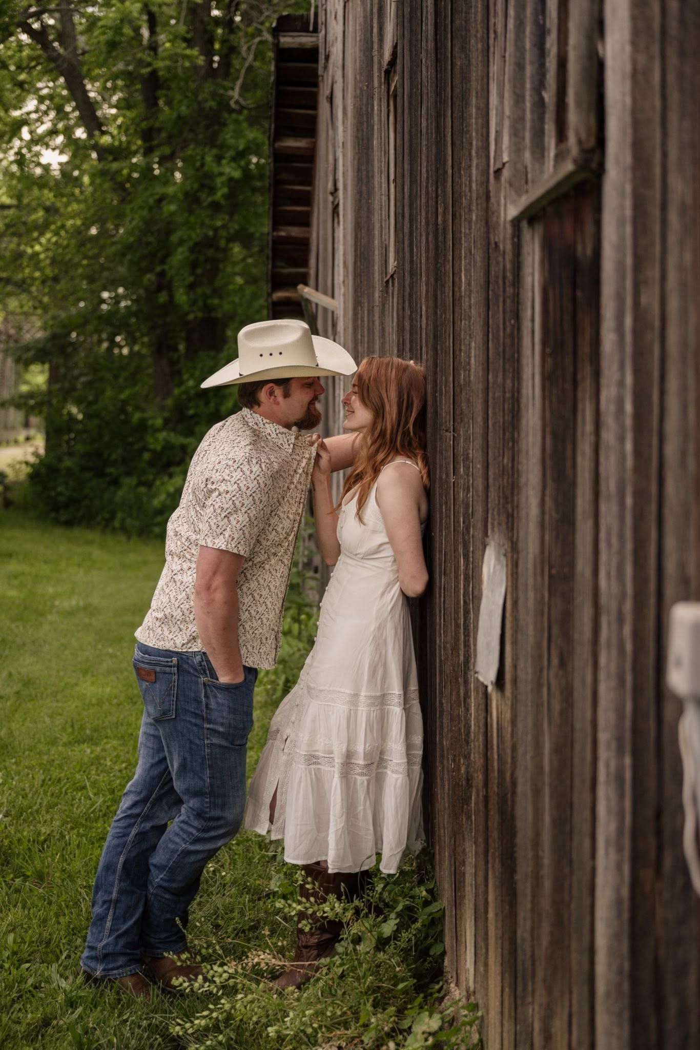 A cowboy in a white hat and jeans leans toward a woman in a white dress against a rustic wooden barn.