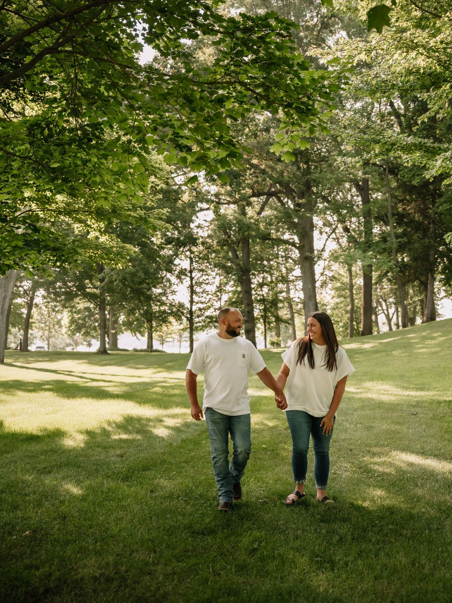 A couple holding hands and walking through a lush green park, both wearing white shirts and jeans on a sunny day.