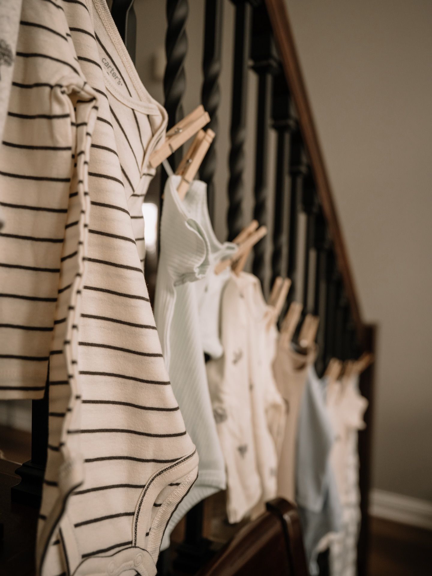 Baby onesies and striped shirts clipped with wooden clothespins along a dark wooden staircase railing.