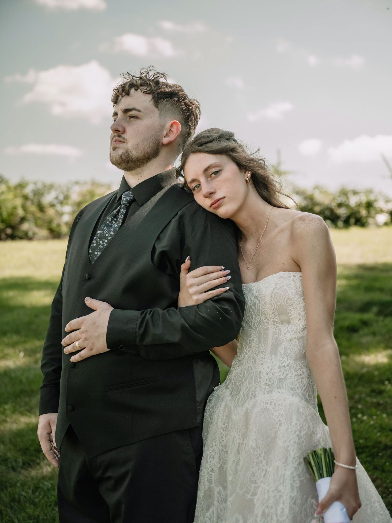 Bride in strapless lace gown leans on groom in black suit outdoors on green lawn under cloudy sky.