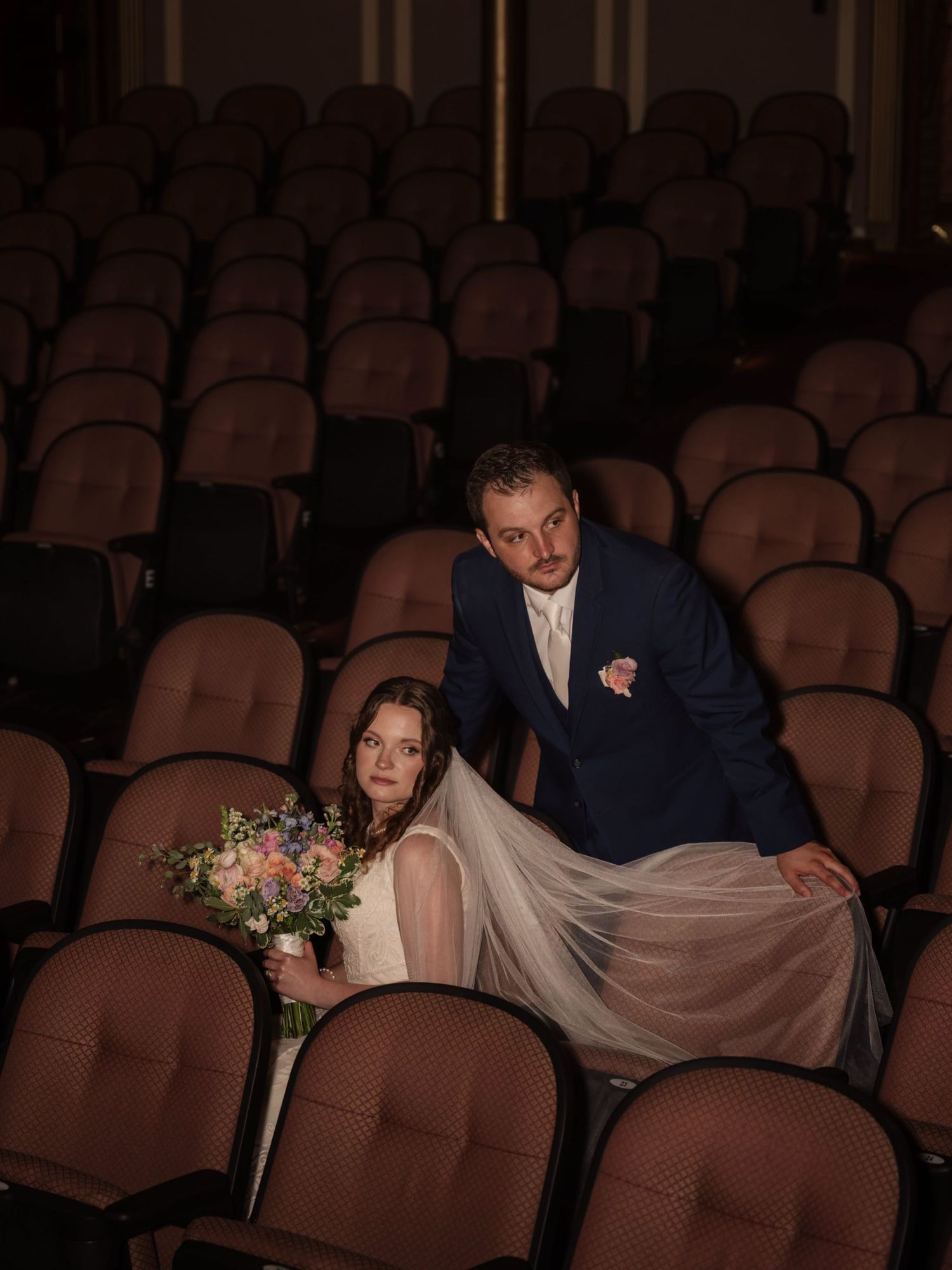Bride and groom pose elegantly among rows of theater seats, bride holding colorful bouquet with flowing veil.