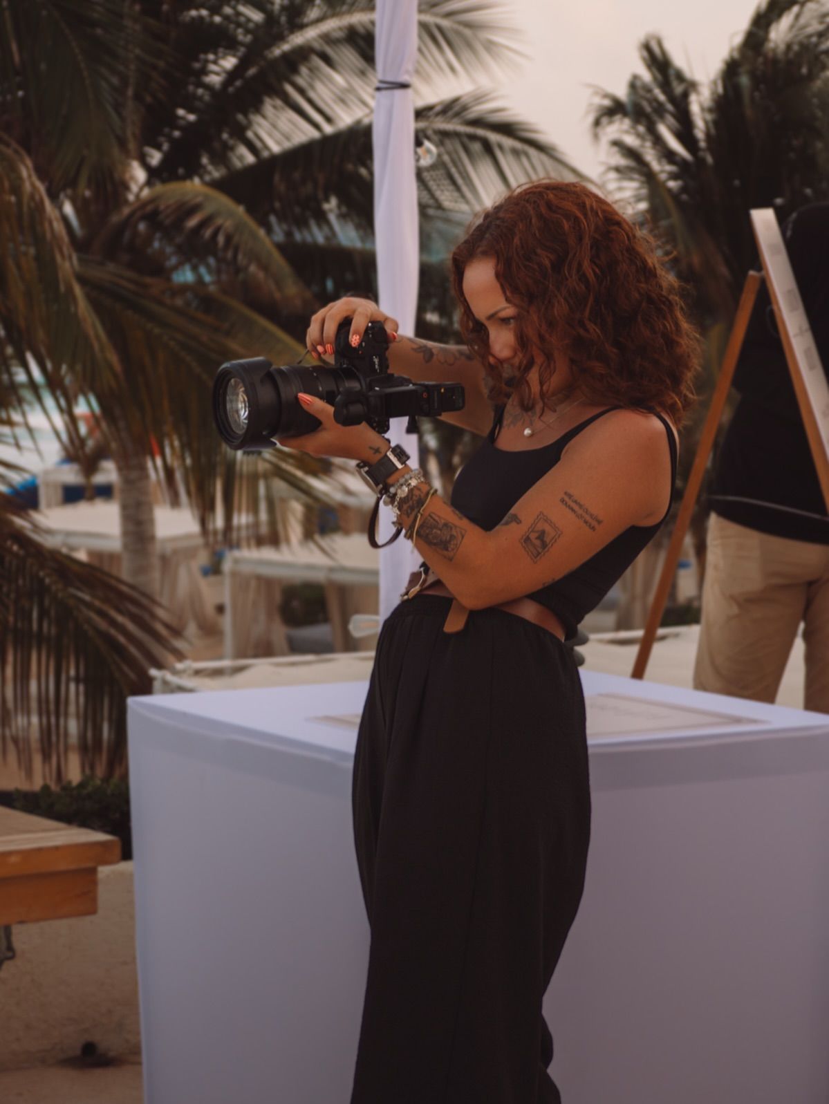 A photographer in black attire working with professional camera equipment among palm trees.