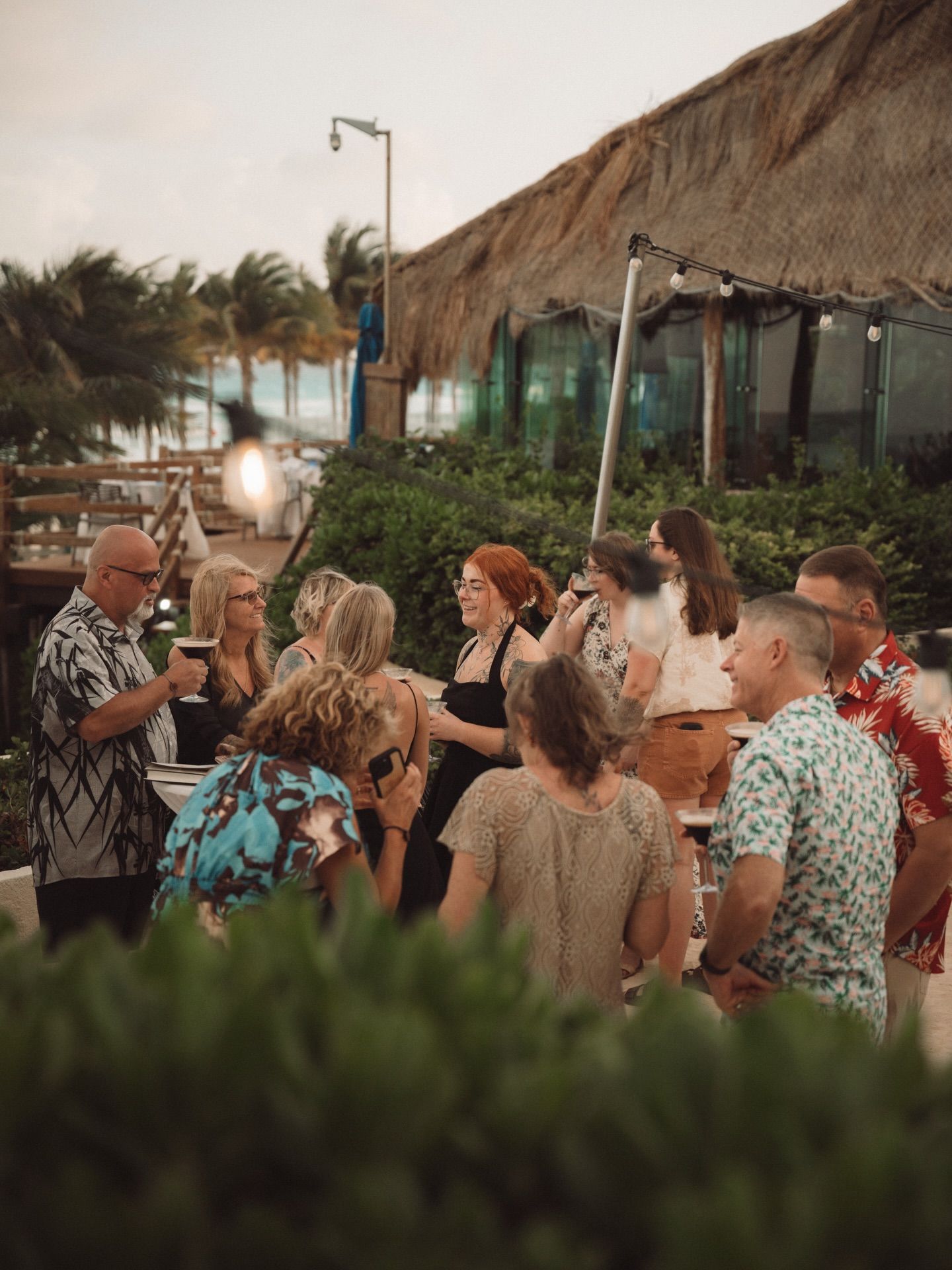 A group of people in tropical attire gather at a beach resort during sunset, creating a festive atmosphere.