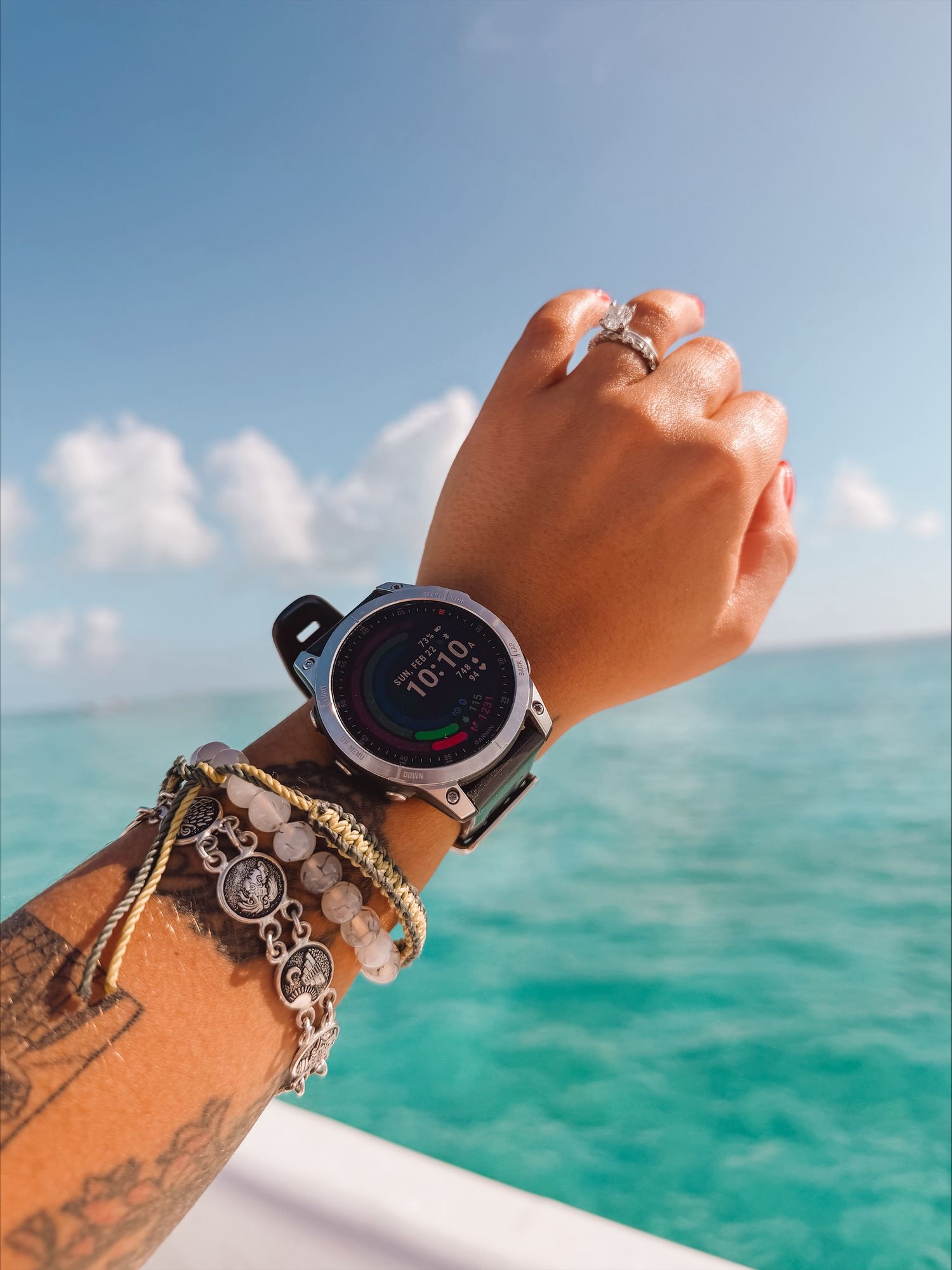 Arm wearing digital smartwatch and bracelets against turquoise ocean water and blue sky with white clouds.