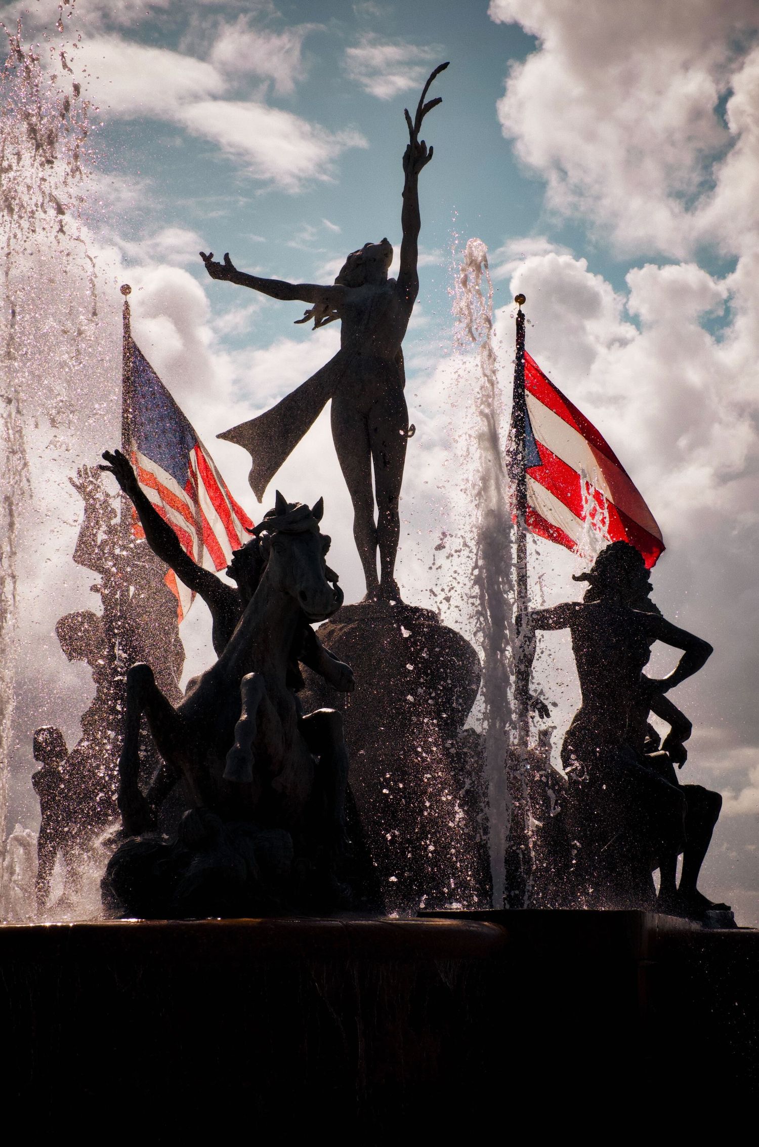 Statue of a woman with outstretched arms on a fountain in Old San Juan with American and Puerto Rican flags in background.