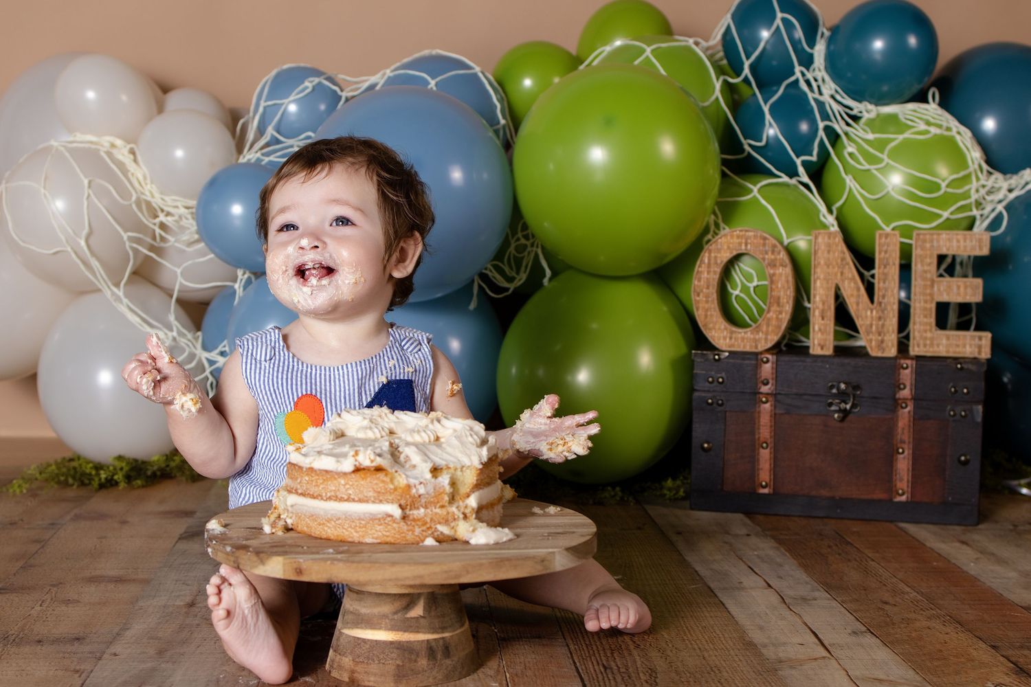 Baby smashing first birthday cake surrounded by green and blue balloons with ONE sign backdrop.