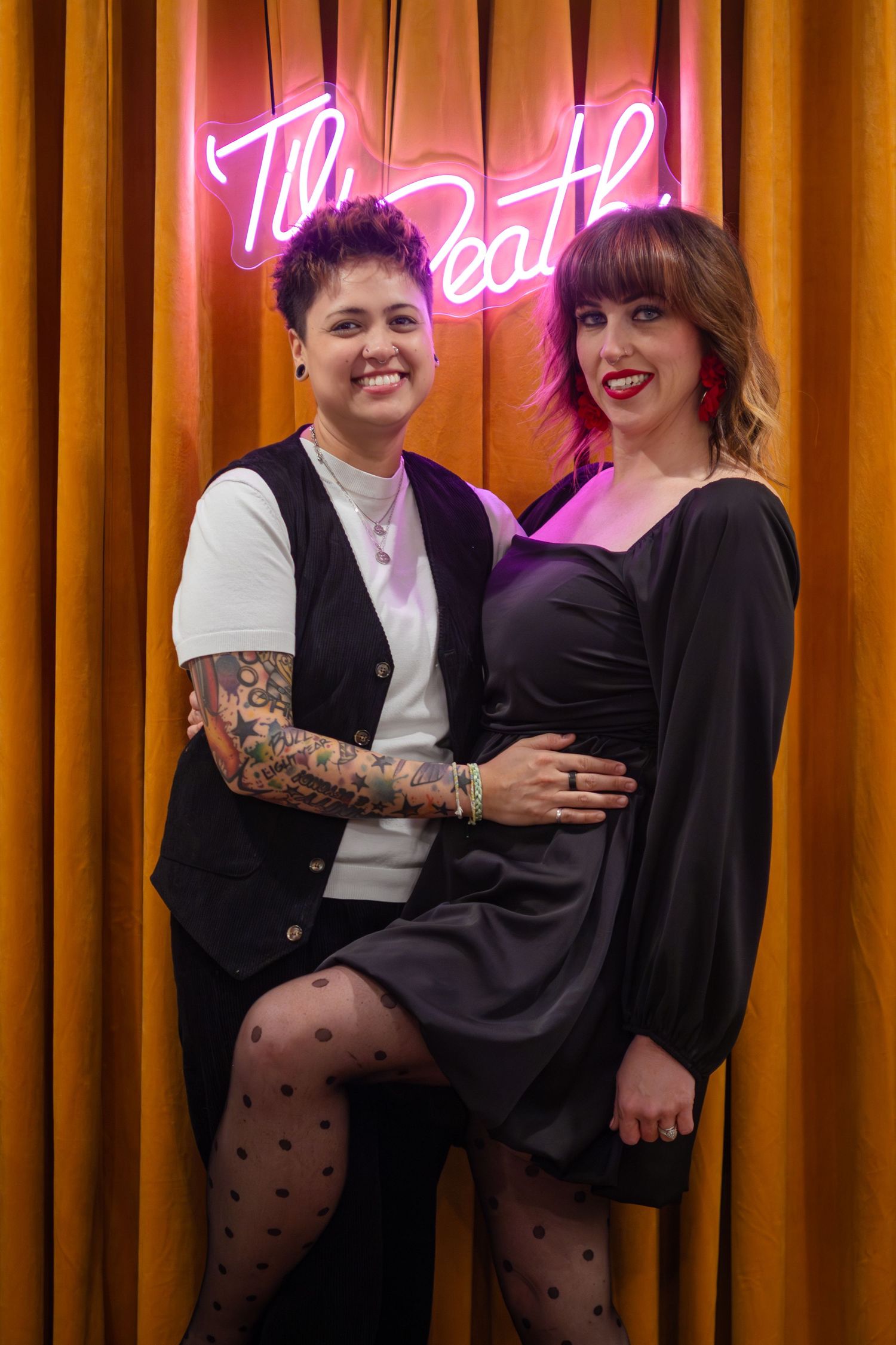 A group of people wearing black outfits pose together in front of orange curtains with a neon sign glowing pink behind them.