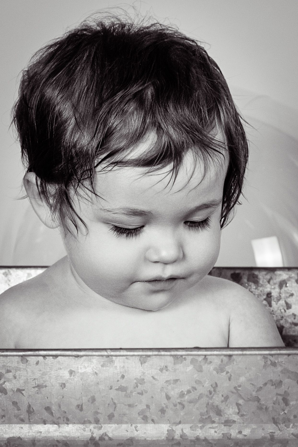 Black and white photo of a cute baby with wet hair sitting in a metal tub, looking down curiously.