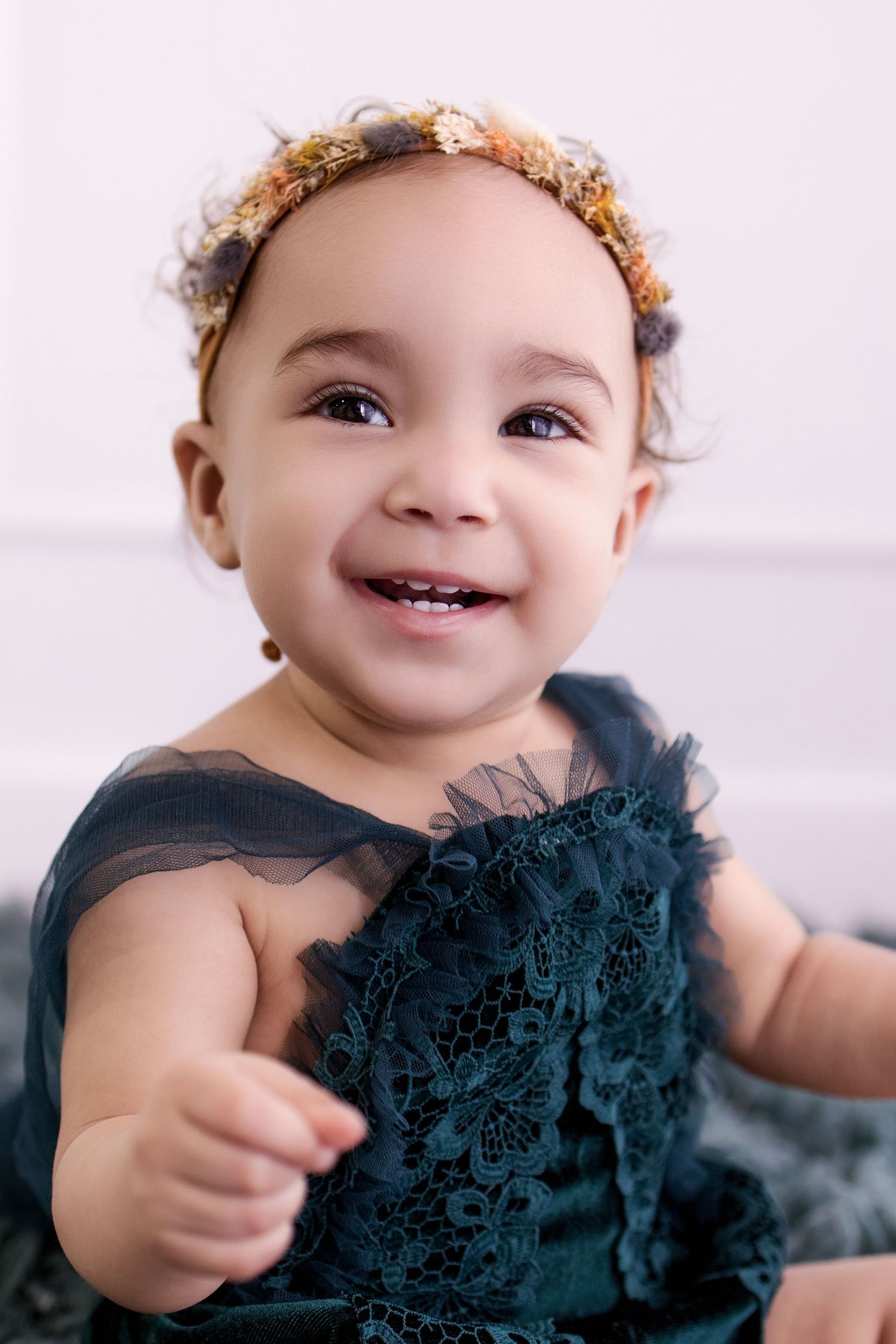 Smiling baby girl in teal lace dress and floral headband, showing tiny teeth in a bright studio portrait.
