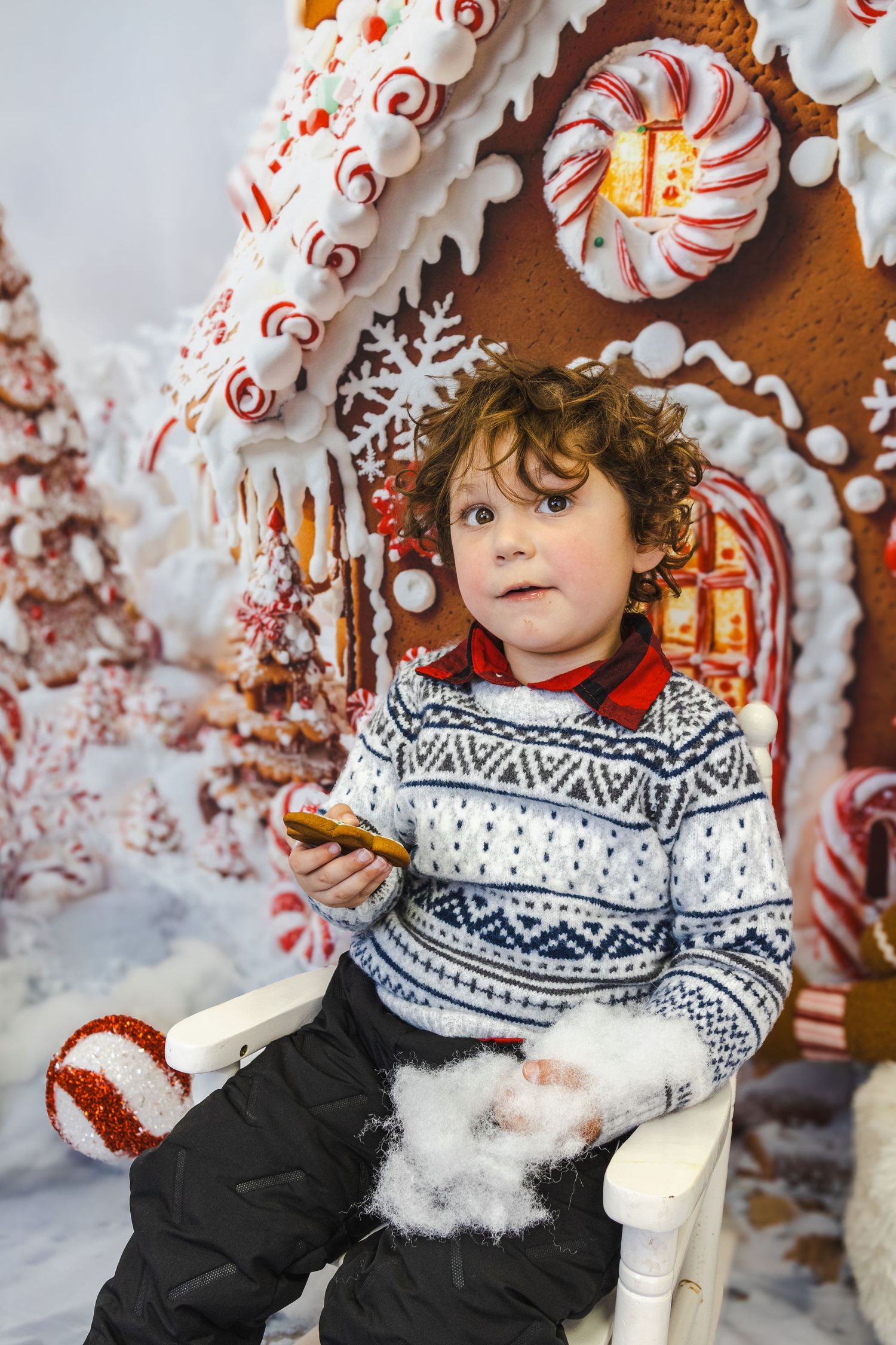 Young boy in Fair Isle sweater holds cookie beside festive gingerbread house with peppermint decorations.