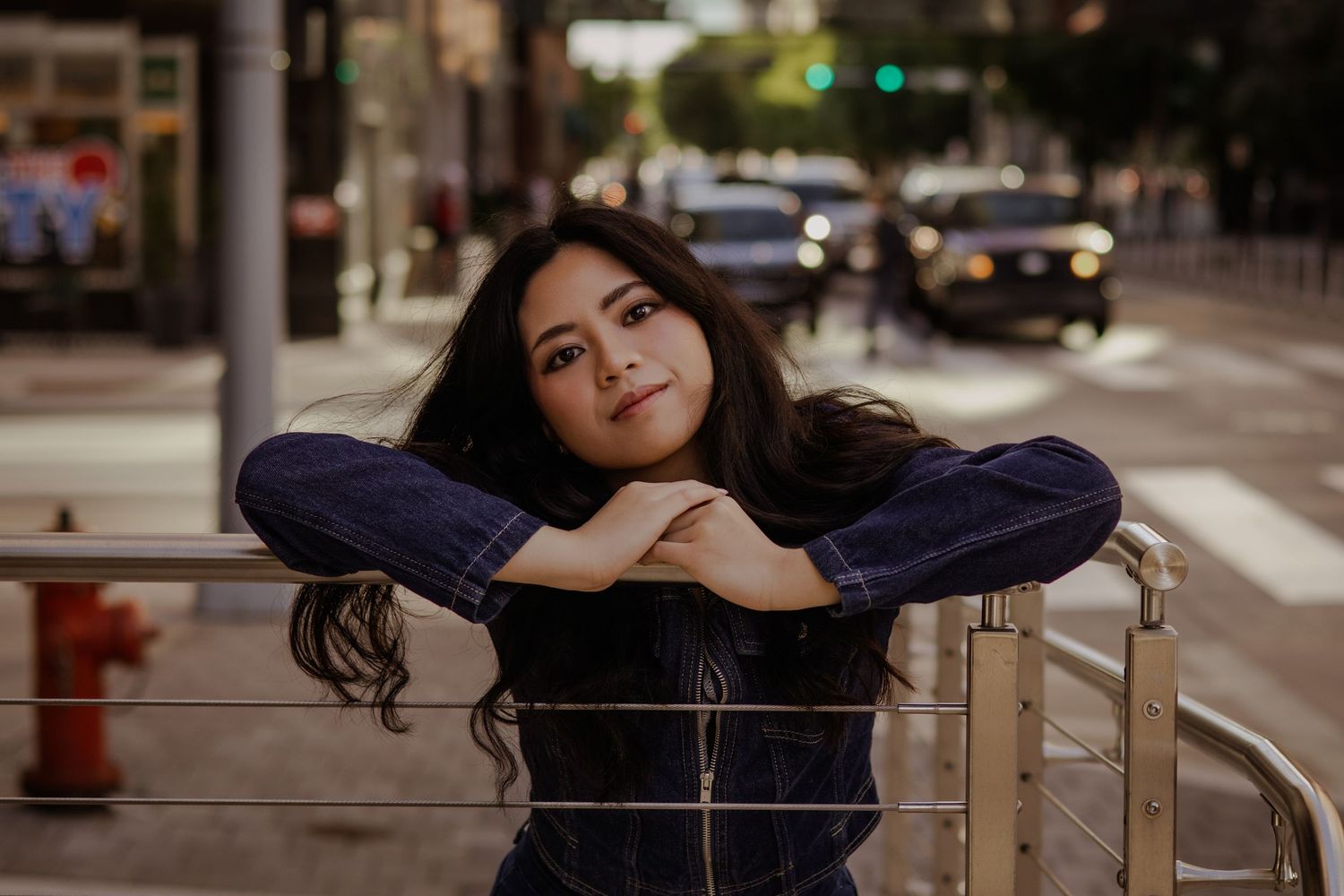 Young woman leans on metal railing on city street, gazing upward with soft bokeh background.