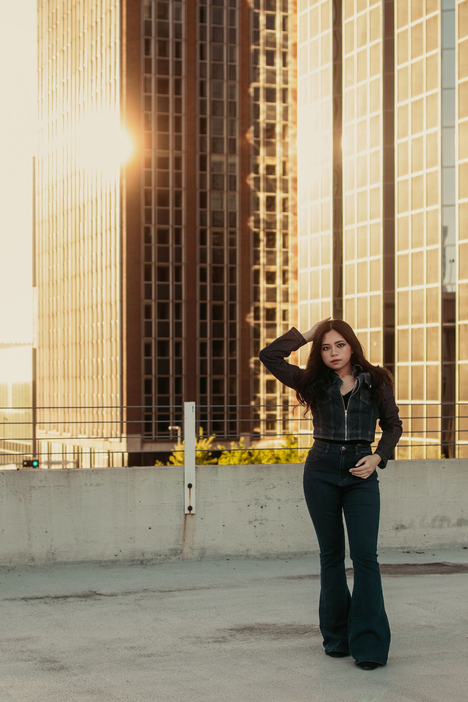 Young woman in black flare jeans poses on rooftop parking with golden sunset reflecting off city skyscraper.