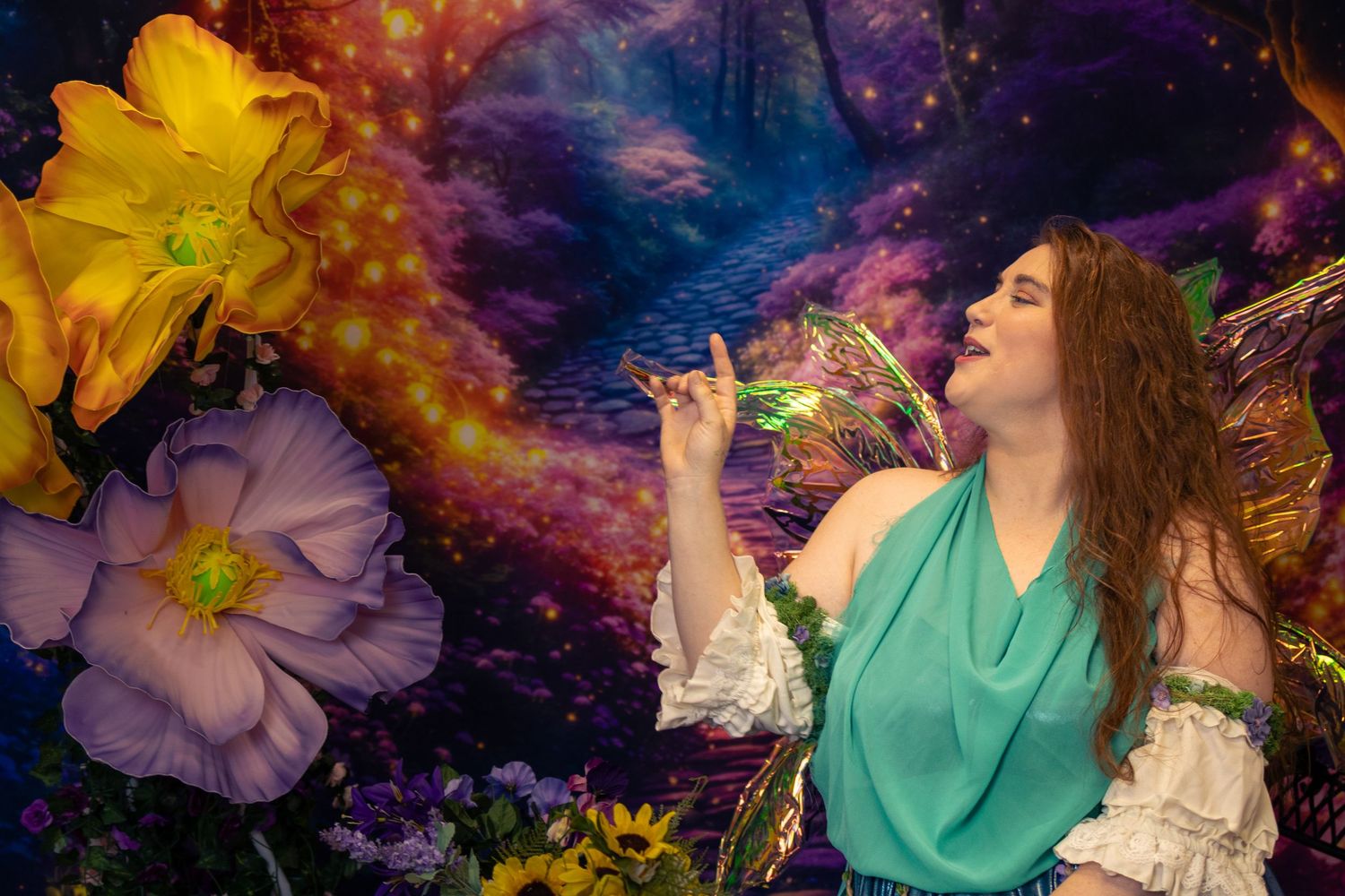 A woman in a teal fairy costume poses among colorful flowers against a magical fantasy forest backdrop.
